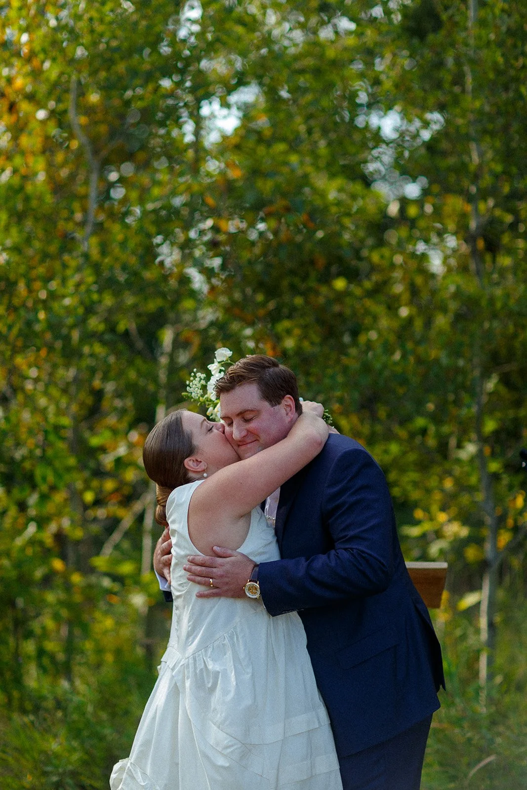 A bride and groom hugging outdoors surrounded by green trees during a wedding ceremony.