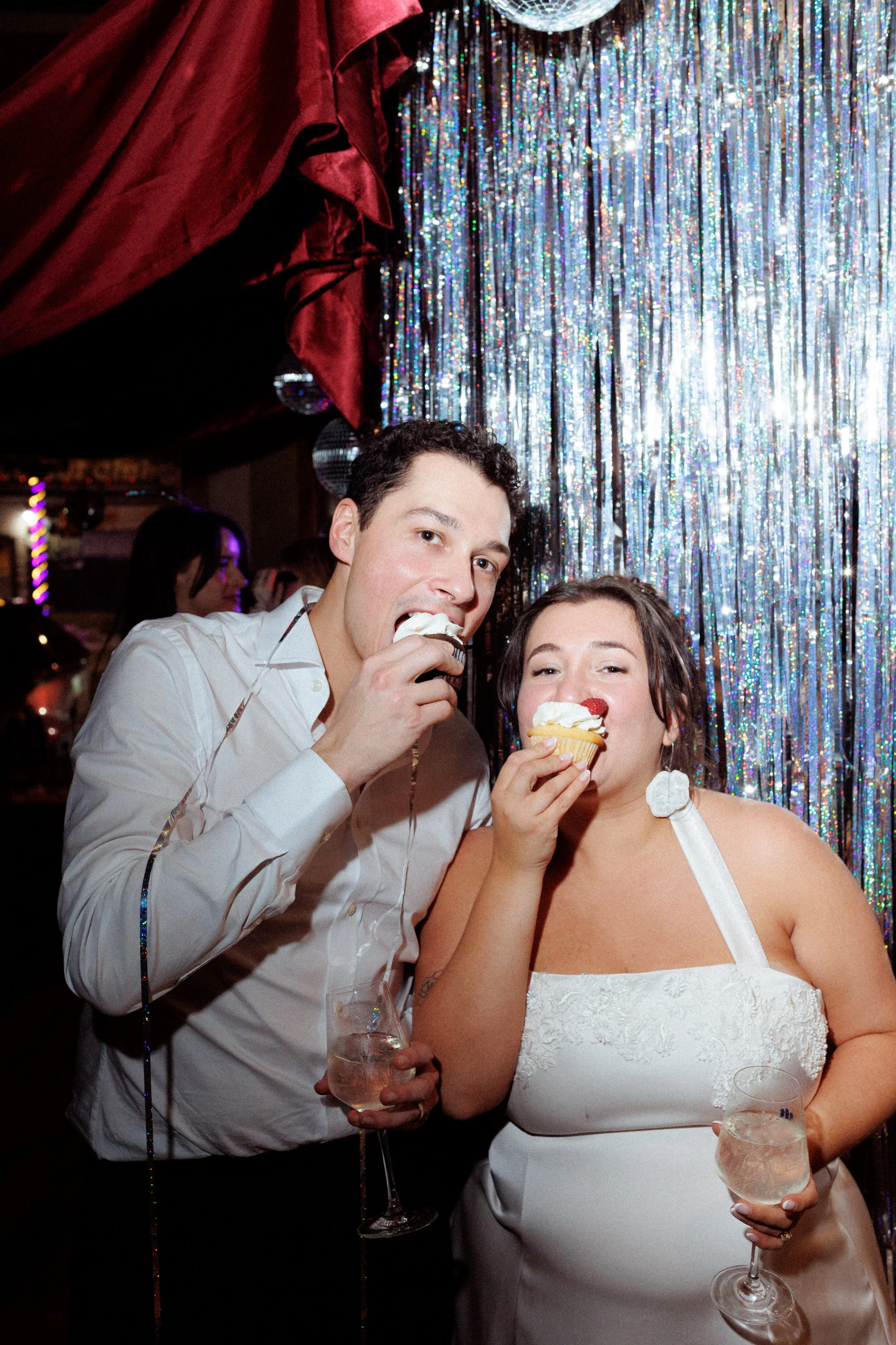 Two people eating cupcakes at a celebration, with a colorful, shiny backdrop and disco ball overhead.