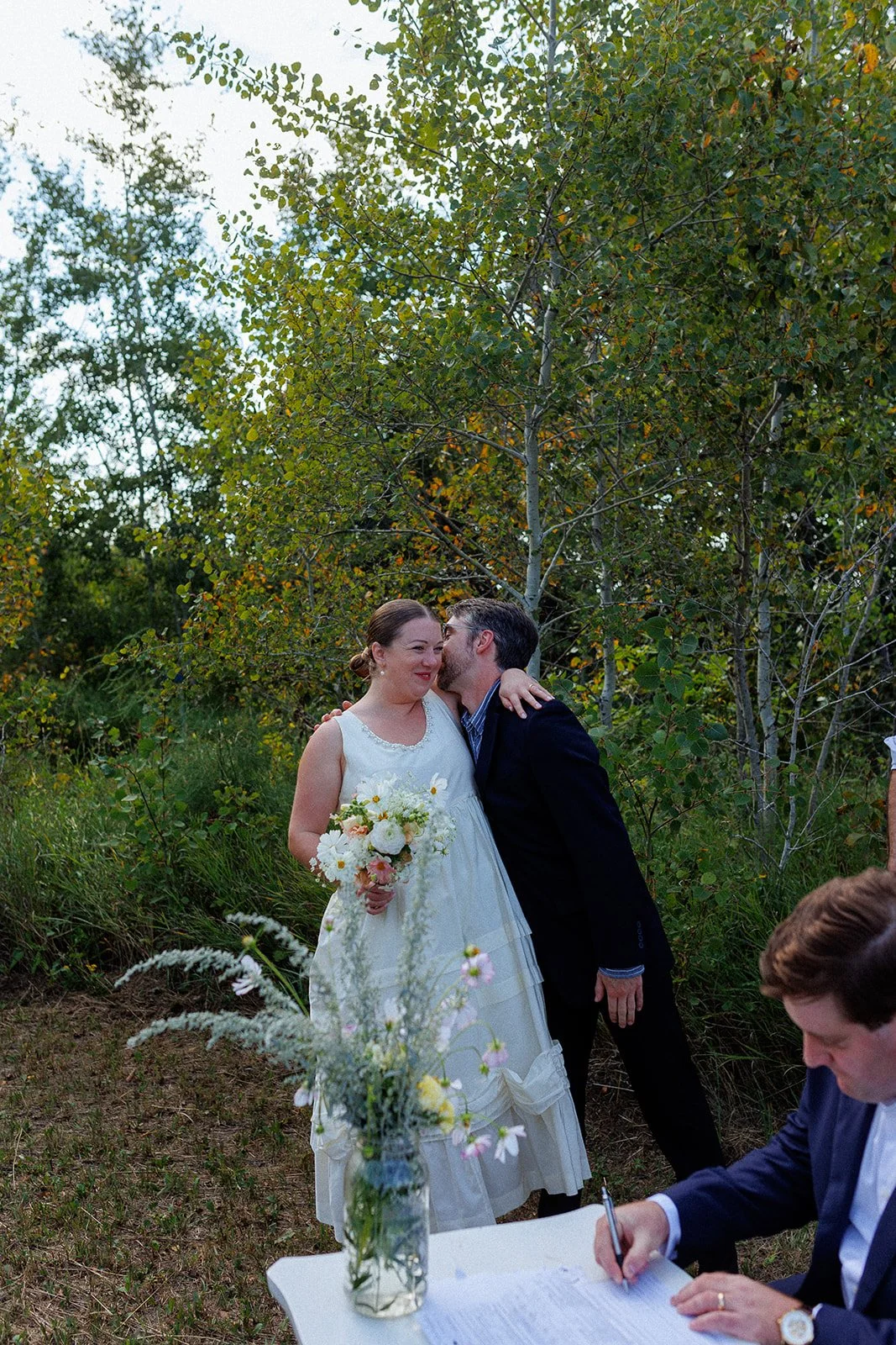 A couple at a wedding ceremony outdoors, with the groom whispering into the bride's ear. The bride is holding a bouquet and wearing a white dress, while the groom is in a dark suit. There is a person sitting at a table signing documents and a vase wi