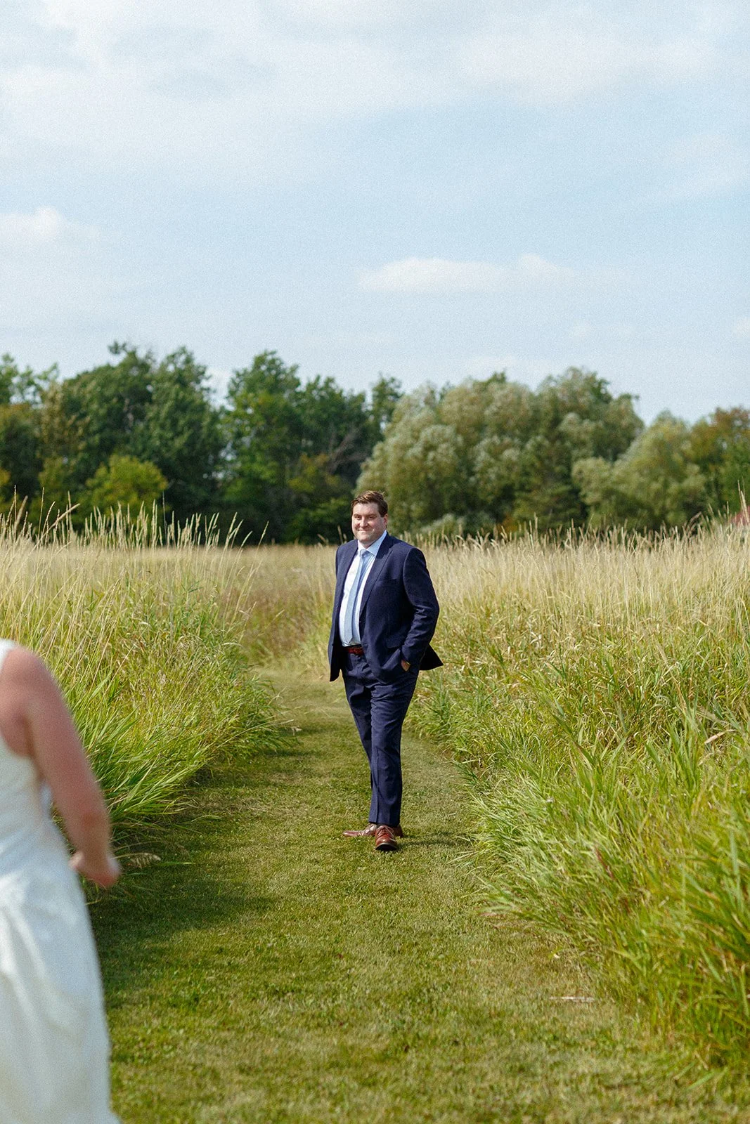 A man in a navy blue suit walking on a grassy path through a field of tall grass, with trees in the background and a partly cloudy sky.