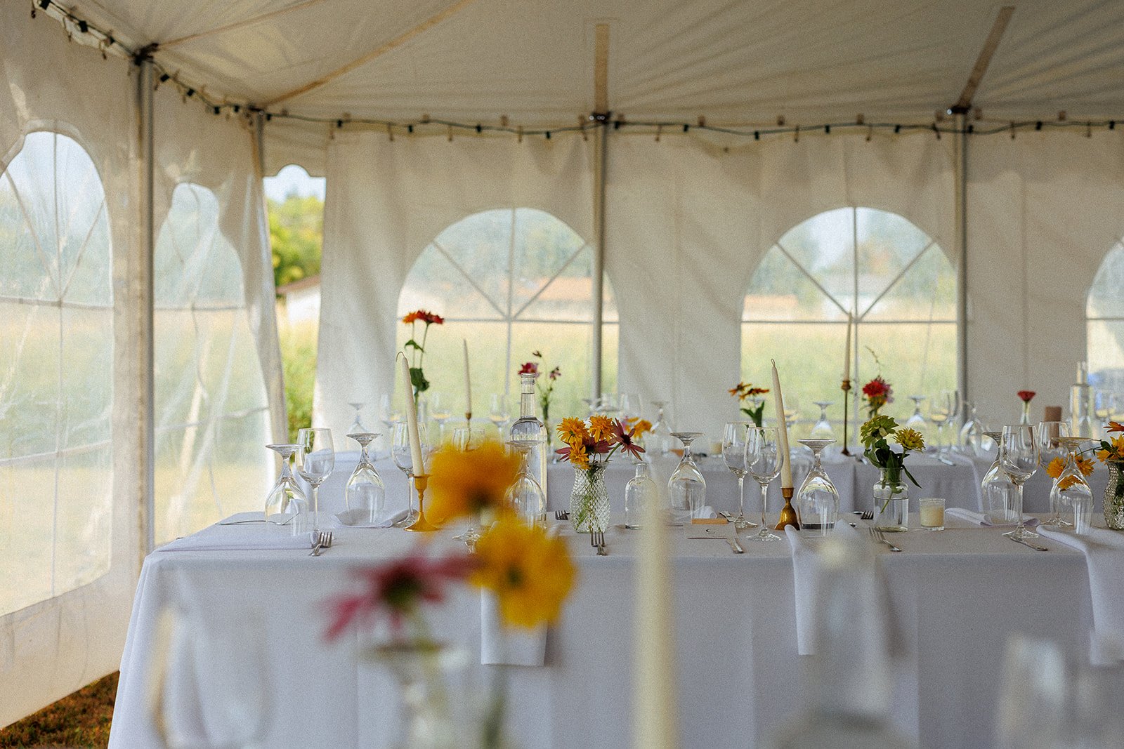 A decorated event tent with a long table set with glassware, flowers in vases, and candles.