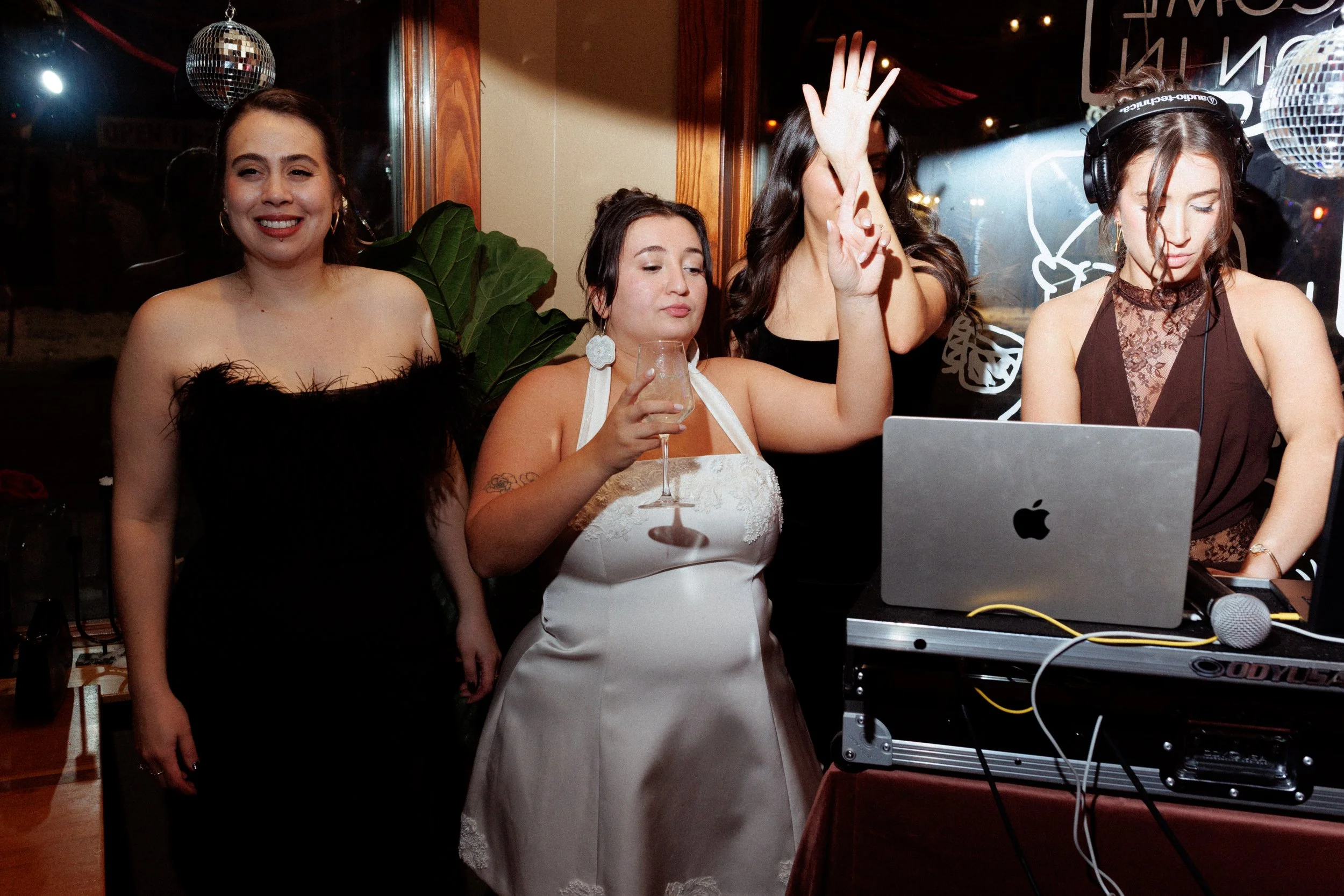 Four women at a party, one wearing a black strapless dress with feather details, another in a white dress holding a wine glass, one with raised hands likely dancing, and a DJ with headphones operating a laptop.