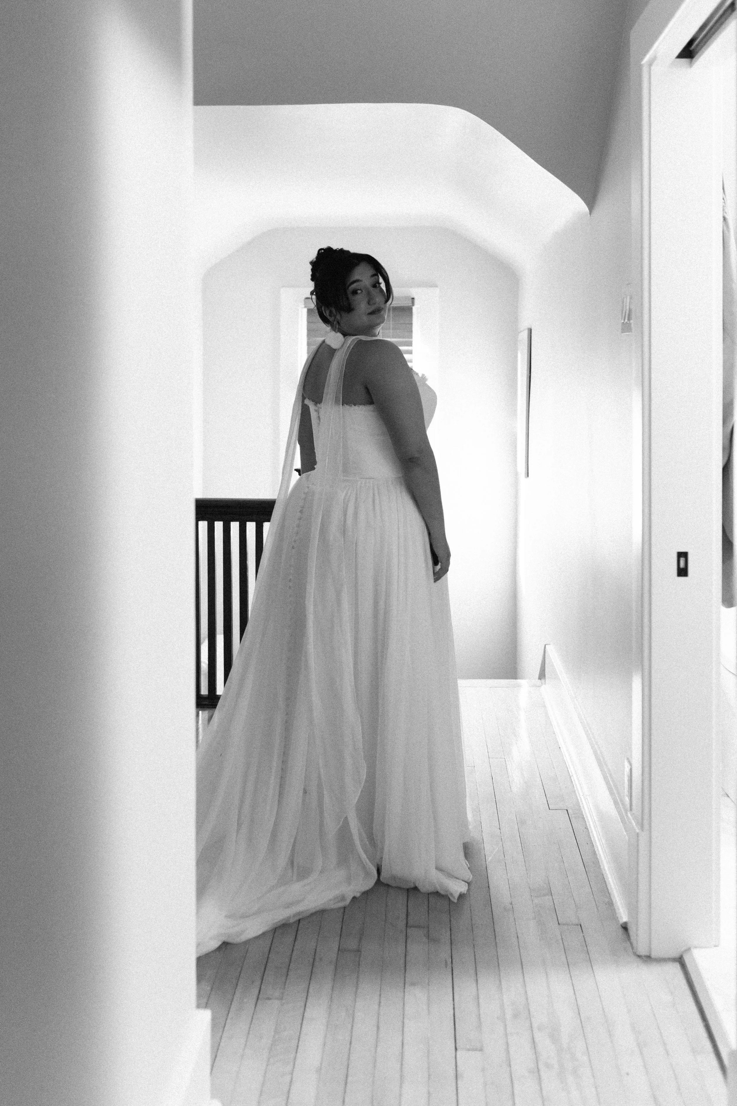 A woman in a white dress standing in a narrow hallway, looking over her shoulder, with a window in the background.