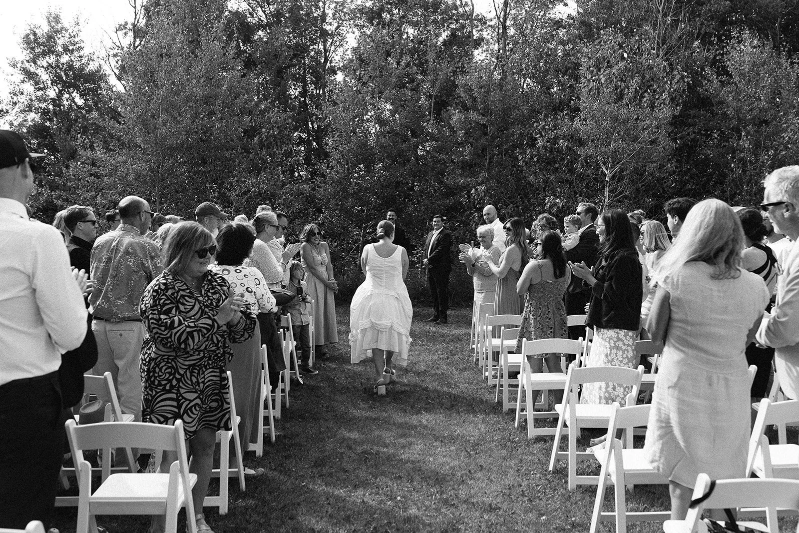 Outdoor wedding ceremony with chairs arranged in two rows, a bride in a white dress walking toward the groom in a suit, surrounded by guests clapping and taking photos, trees in the background.