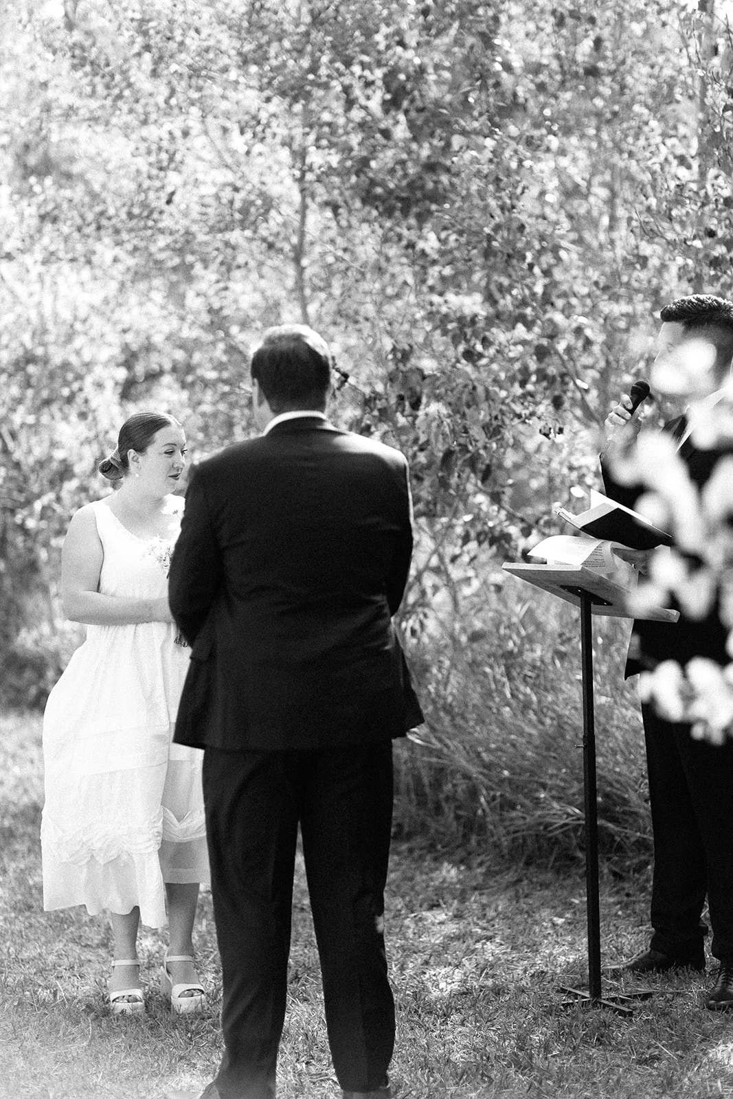 A wedding ceremony outdoors with a woman in a white dress and two men, one in a tuxedo and one reading from a book, in front of trees.