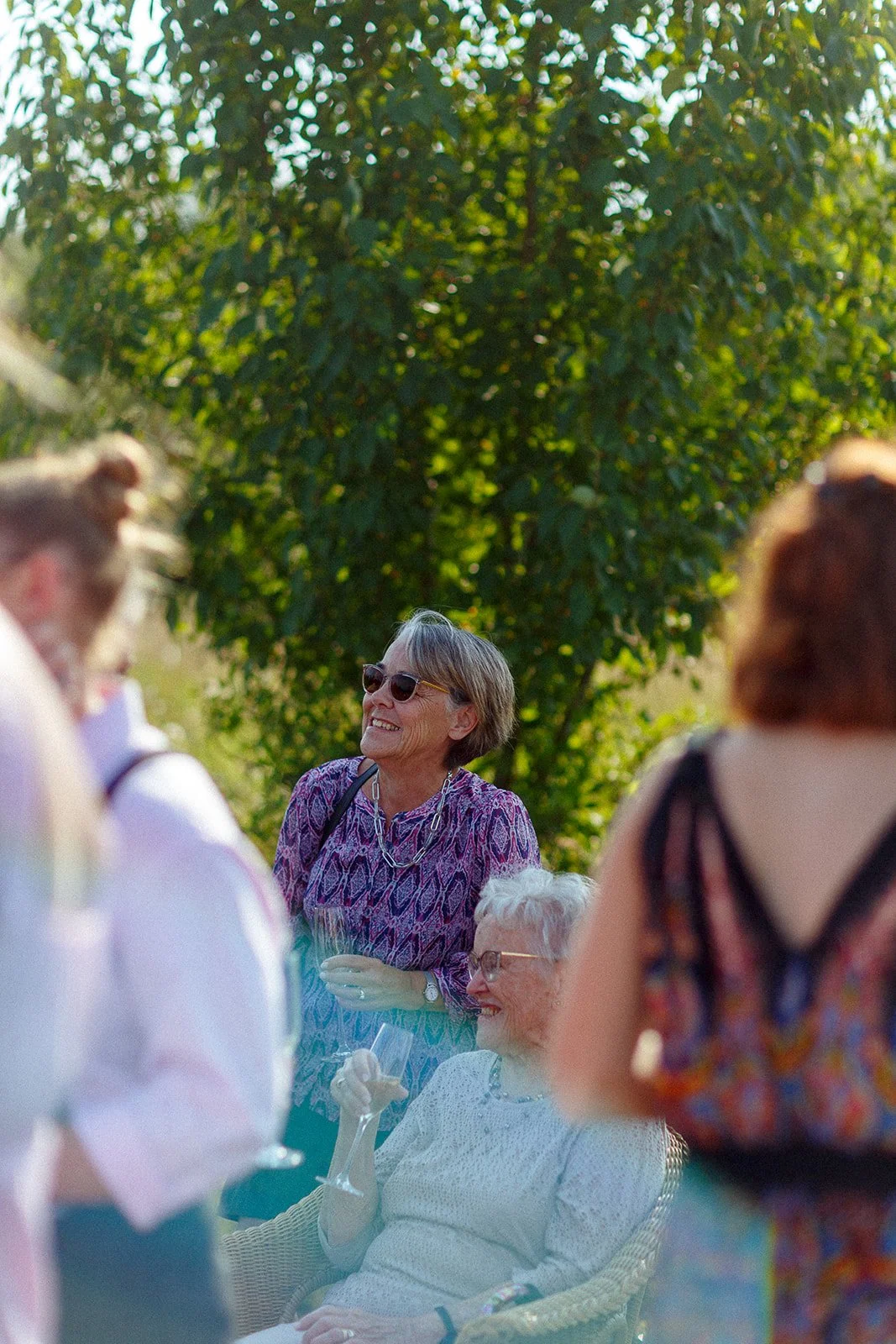 People socializing outdoors with trees in the background, including elderly women smiling and holding drinks, wearing sunglasses and summer clothing.