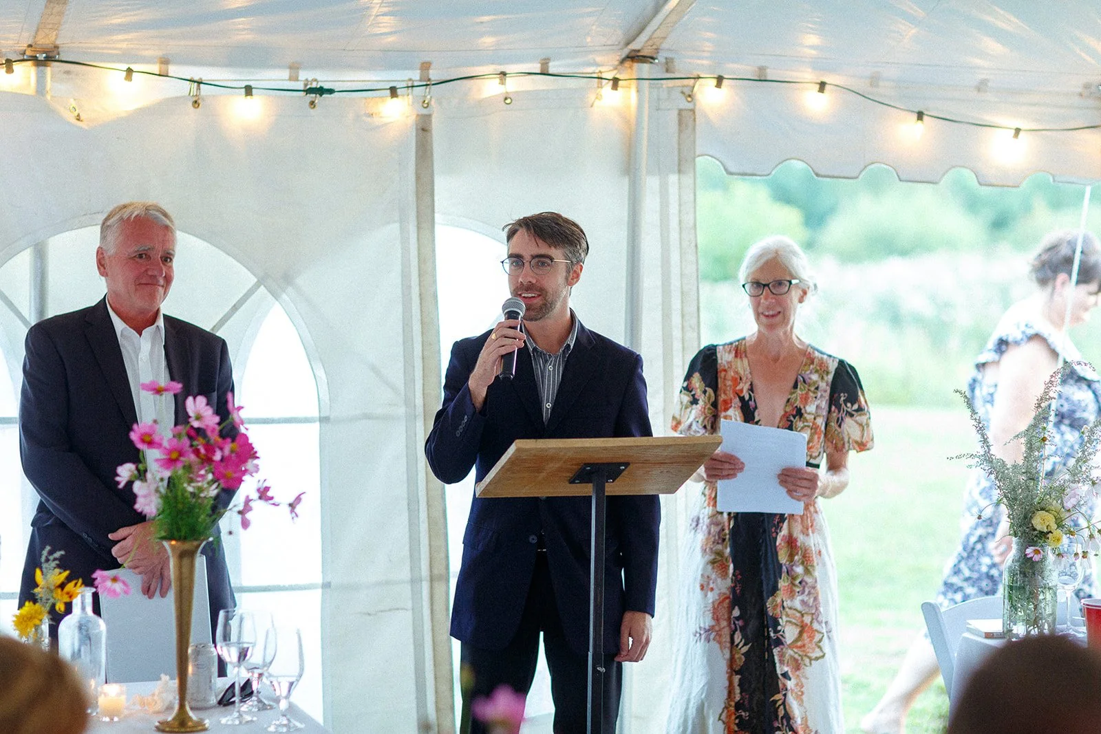 Man in a dark suit giving a speech at a wedding reception, standing behind a wooden podium, with an older woman holding a paper and a man with a flower arrangement in front of him.