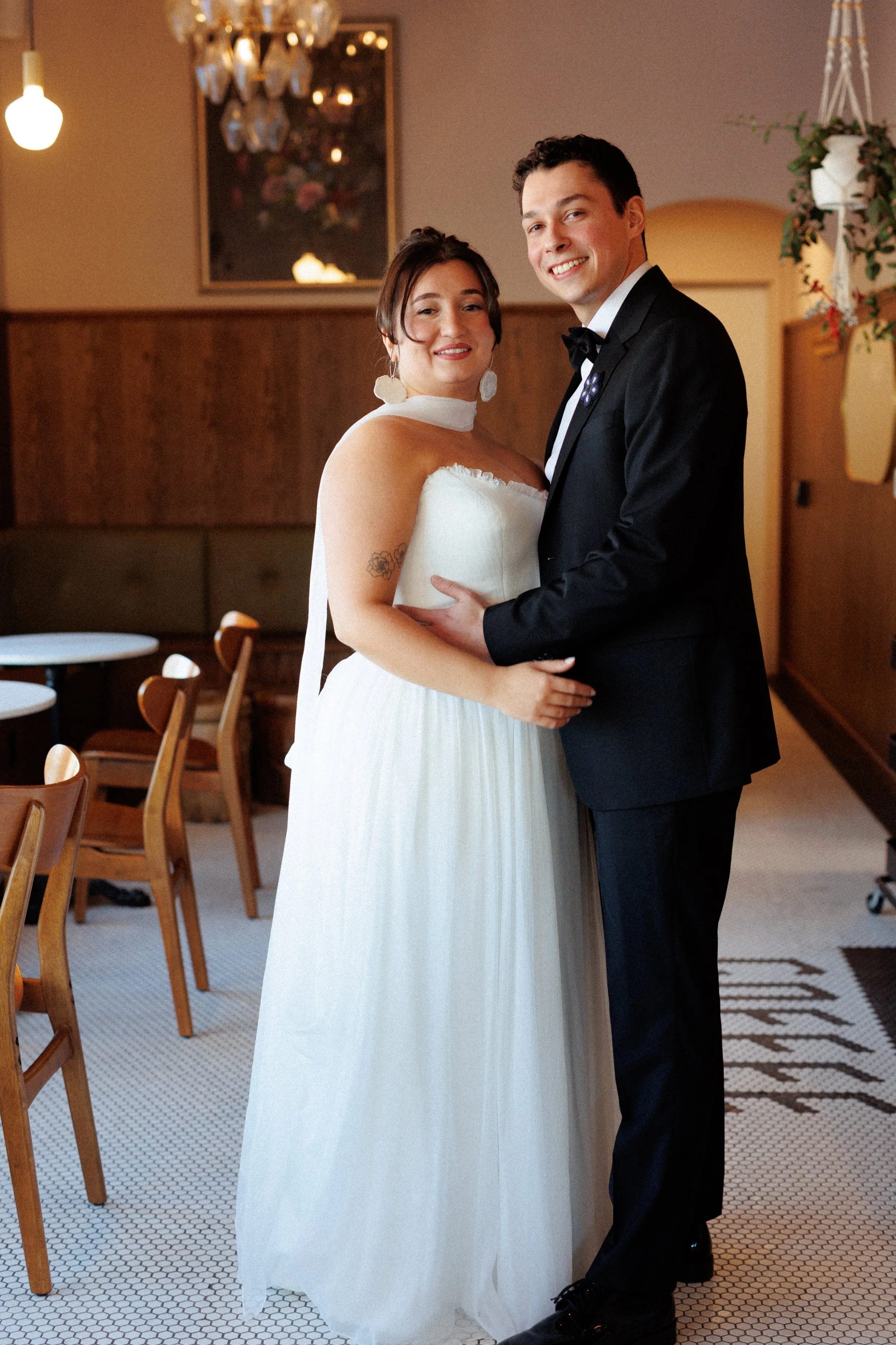 A bride and groom smiling and holding each other in a cozy, warmly lit room, with the bride wearing a white strapless wedding gown and the groom in a black tuxedo with a bow tie.