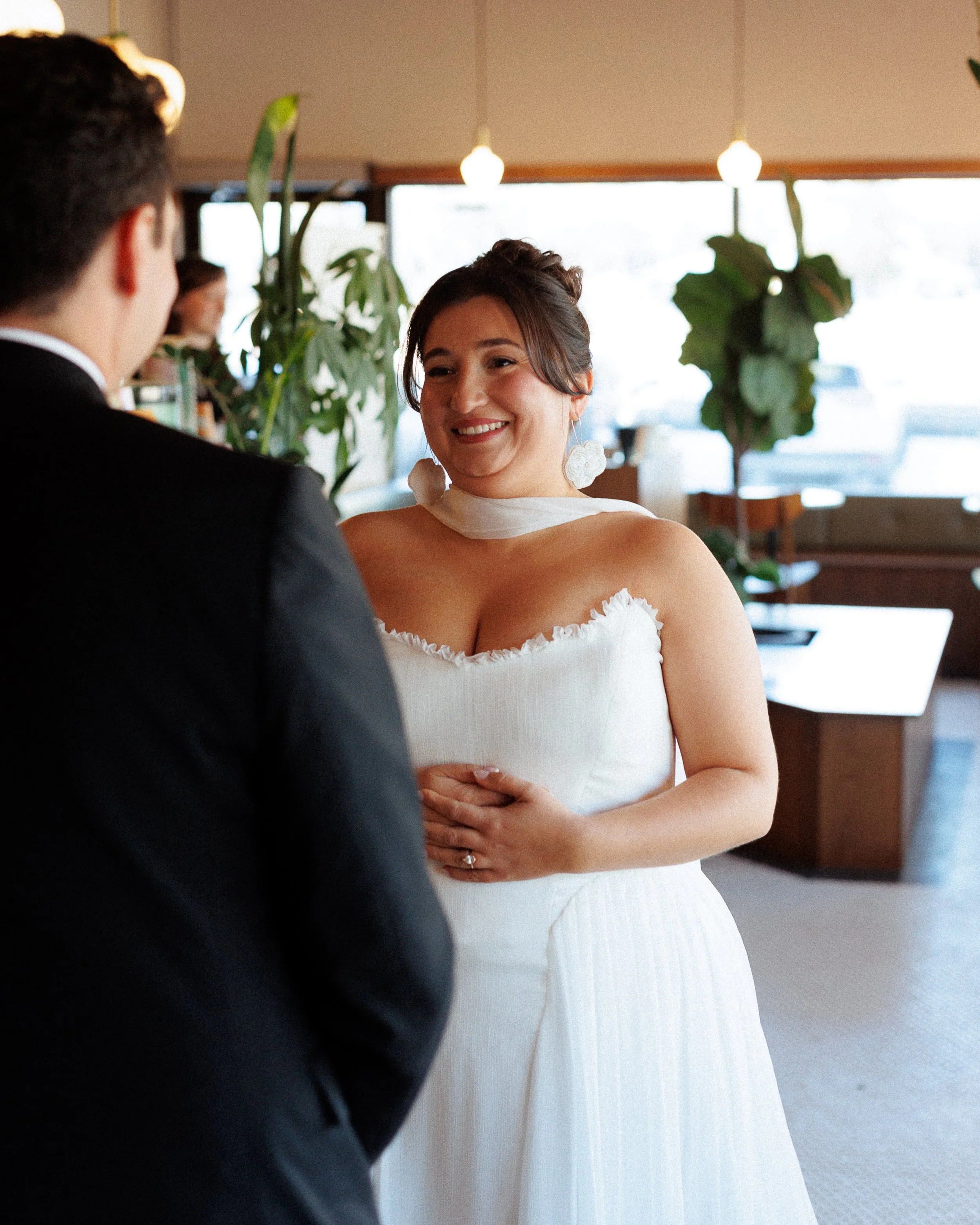 A woman in a wedding dress smiling at a man in a suit during a wedding ceremony inside a warmly lit venue with large windows and plants in the background.