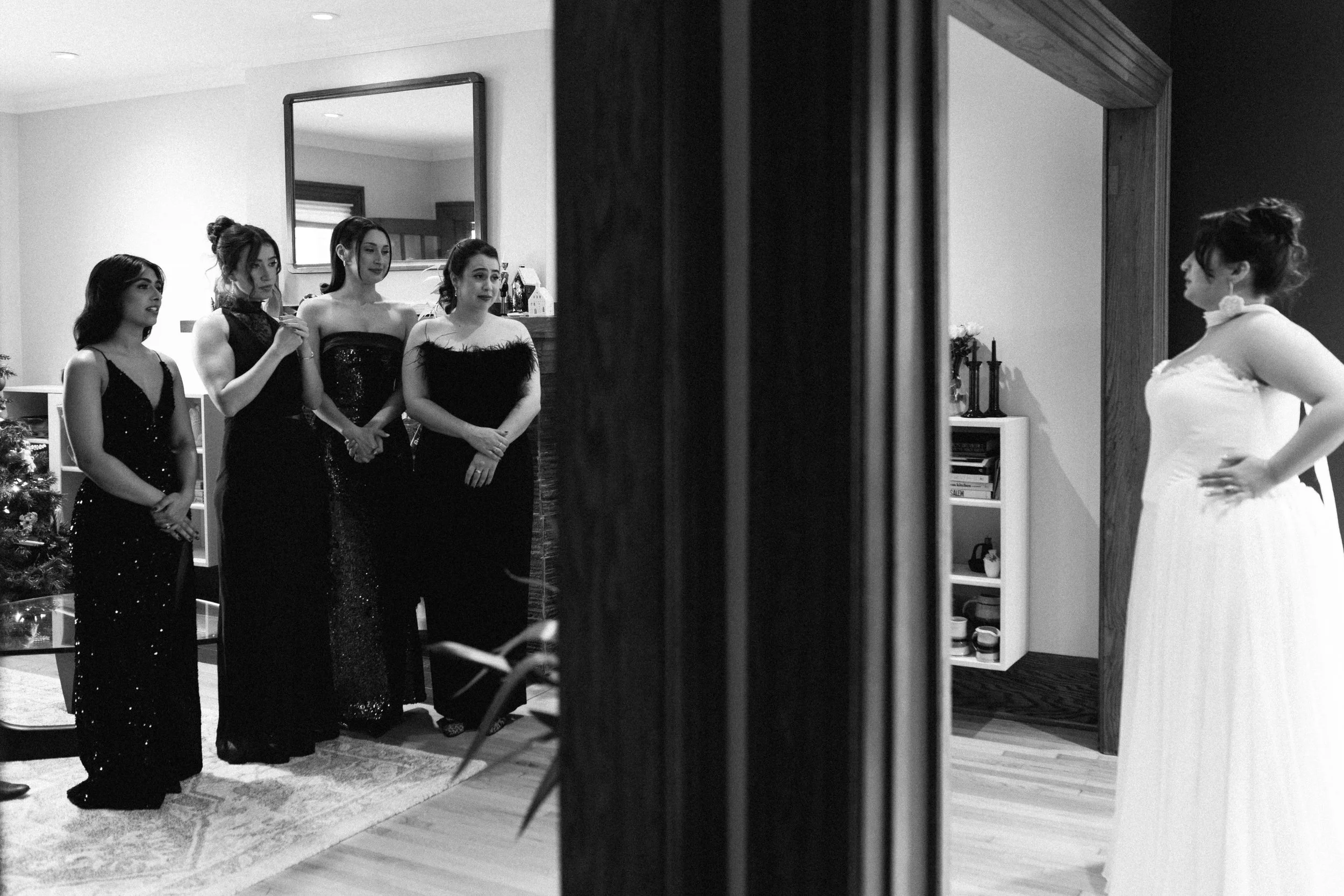 Four women in evening gowns looking at a woman in a white dress, seen through a mirror.