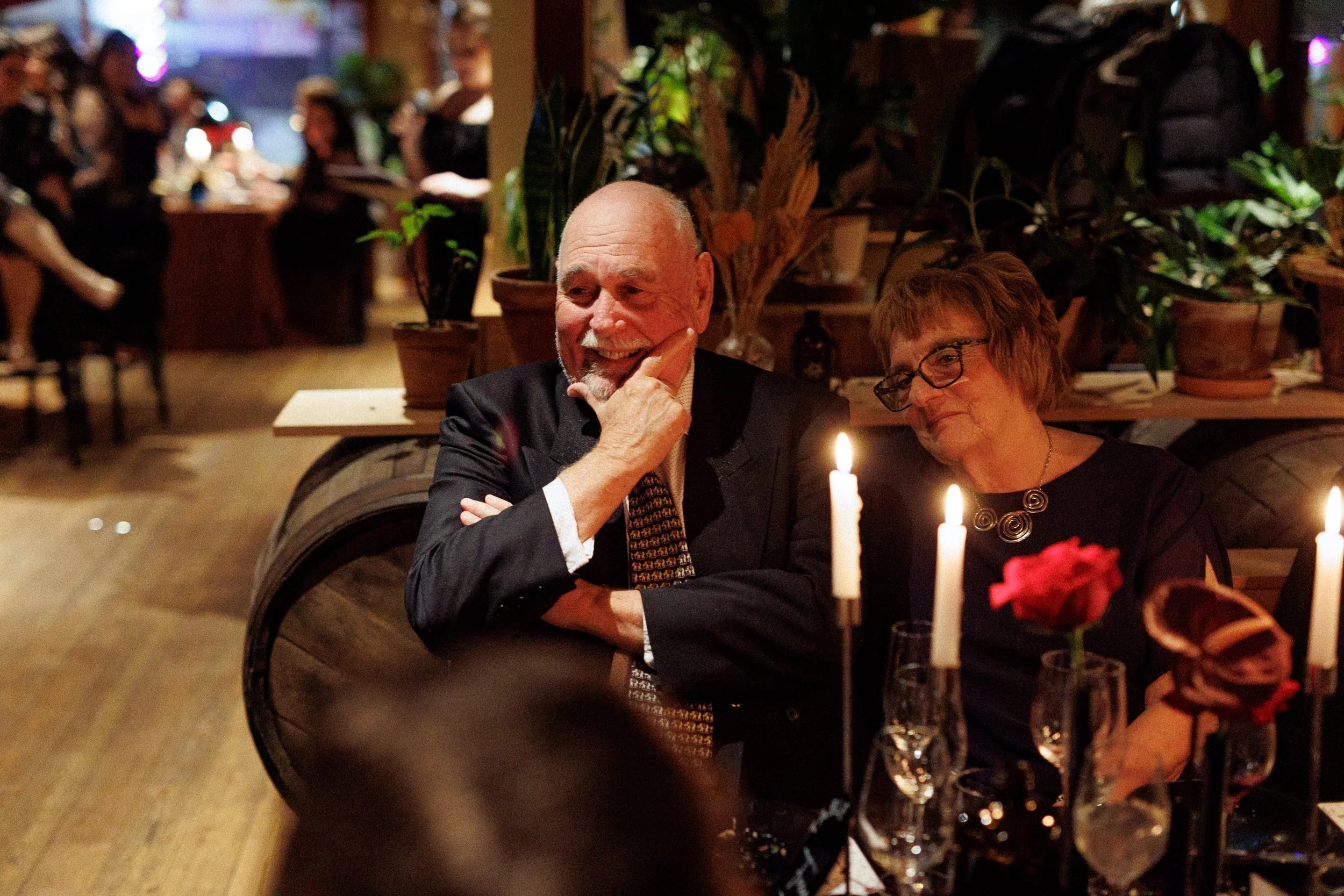 A man and woman sitting at a candlelit dinner table, smiling and engaging in conversation. The man has a beard, is wearing a suit, and appears joyful. The woman has short hair, glasses, and is wearing a dark dress with a necklace. There are candles a