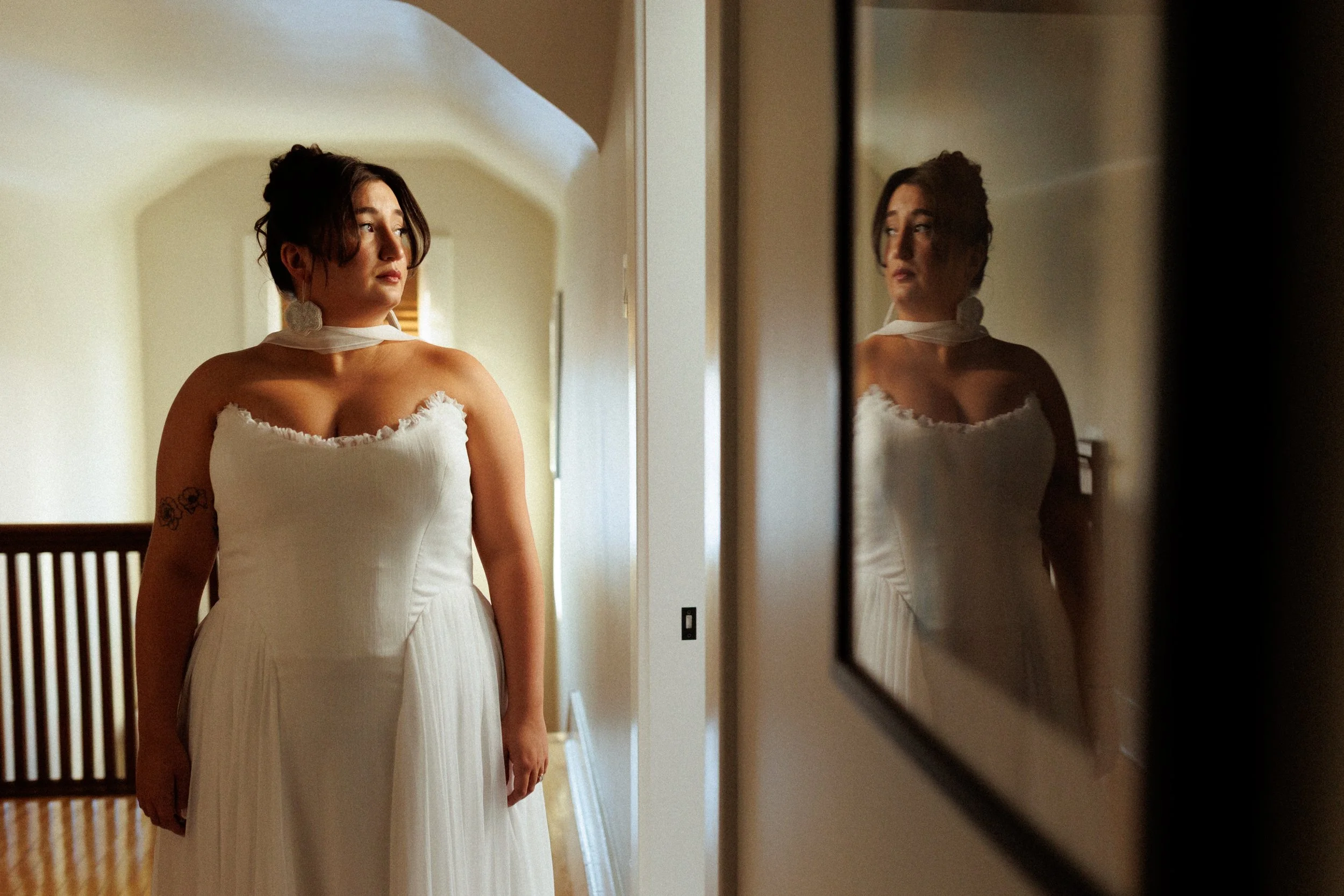 A woman in a white dress standing in front of a mirror, with her reflection visible on the right side of the image.