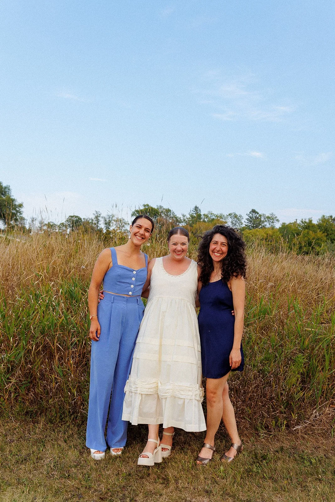 Three women standing outdoors in a grassy field, dressed in summer clothing, smiling at the camera with a clear sky above.