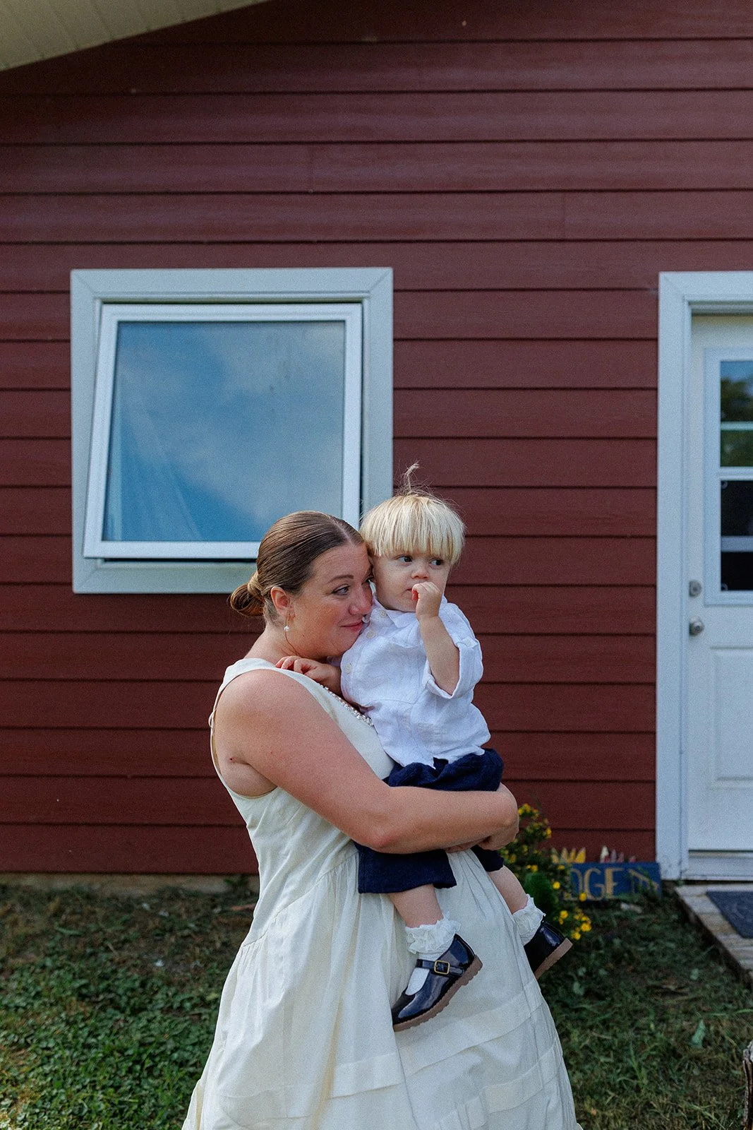 A woman holds a young boy in front of a red wooden house with a slanted window and a white door, outdoors on a grassy area.