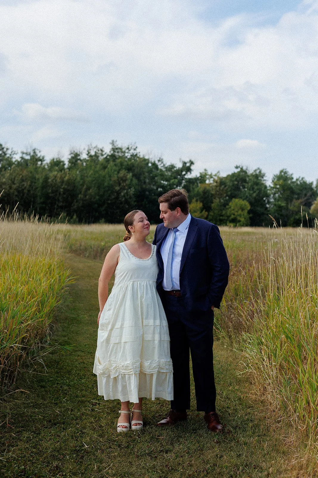 A couple dressed in formal attire standing on a grassy path in a field with tall grass, looking at each other under a partly cloudy sky.