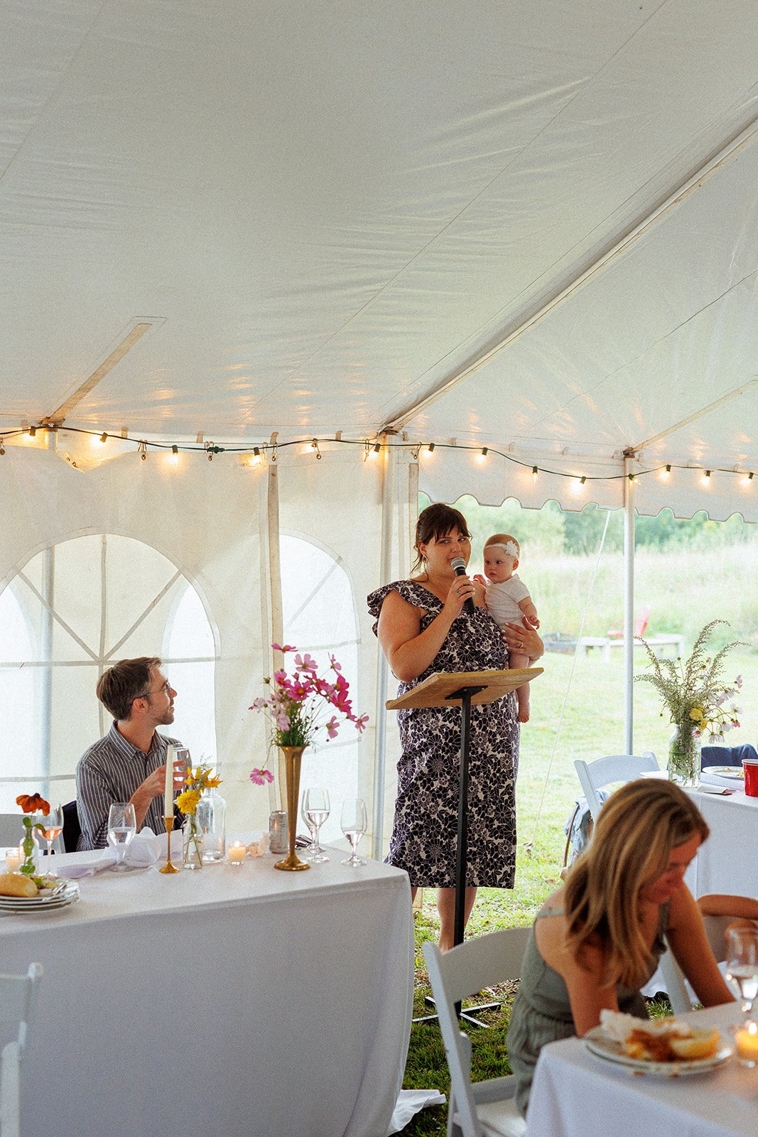 Woman giving speech with a microphone while holding a child in a wedding reception under a tent, decorated with string lights and floral centerpieces.