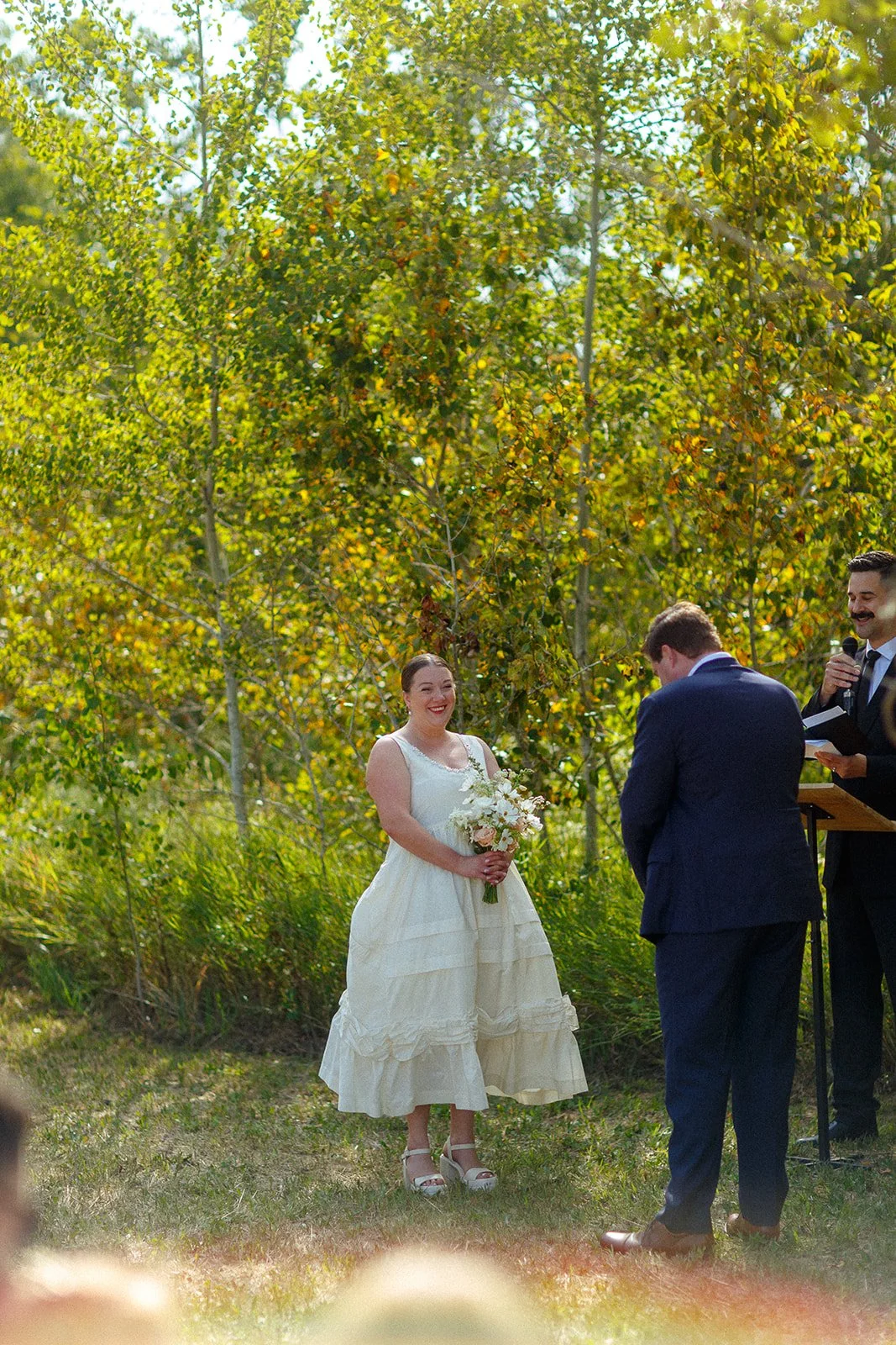 A woman in a white dress holding a bouquet of flowers during a wedding ceremony outdoors, with a man in a dark suit facing her, and an officiant speaking into a microphone.