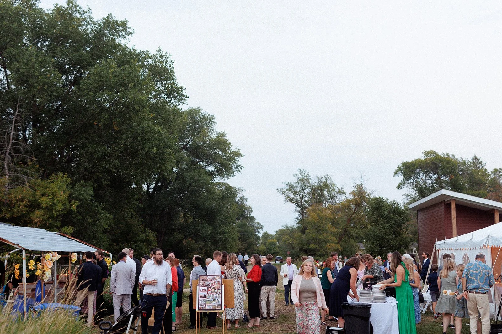 Outdoor wedding reception with guests mingling near decorated tents and tables, set against a backdrop of trees under a cloudy sky.