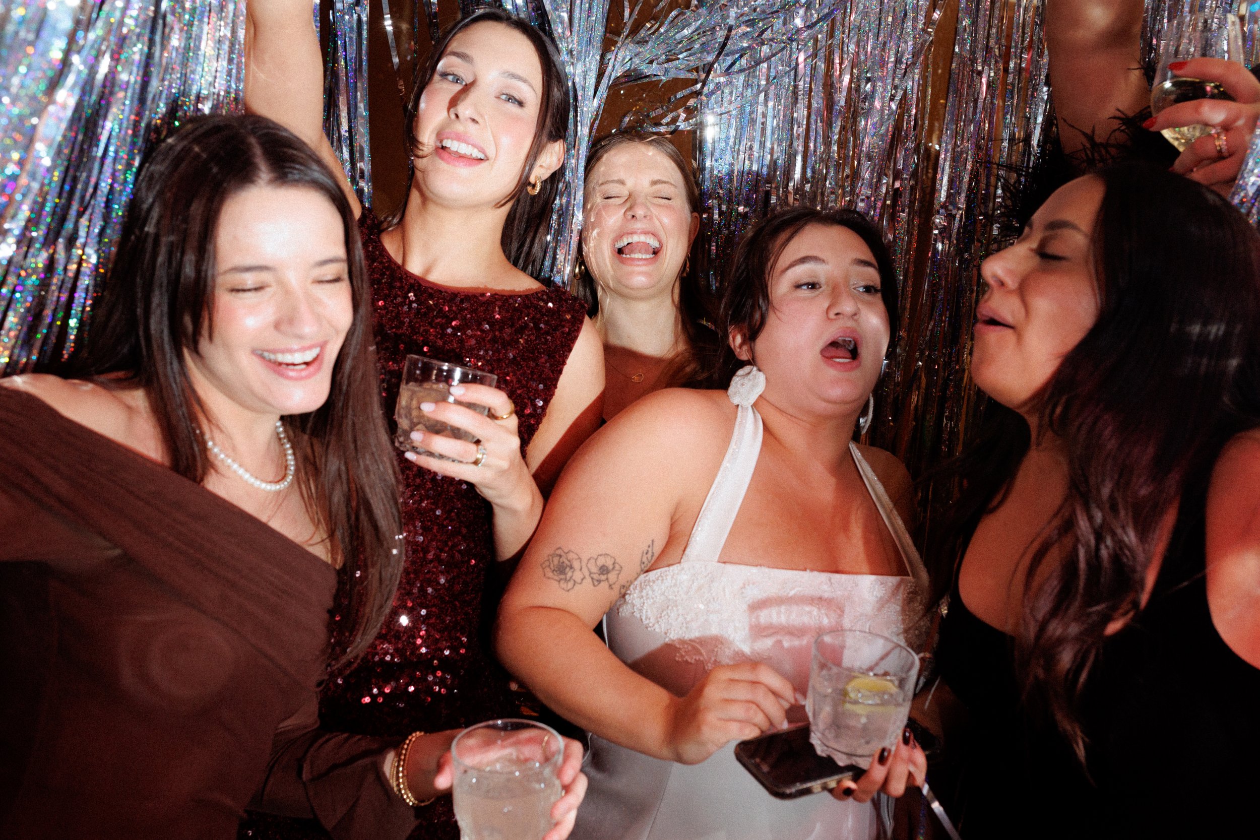 Group of women celebrating at a party, some with glasses of drinks, in front of a sparkly backdrop.