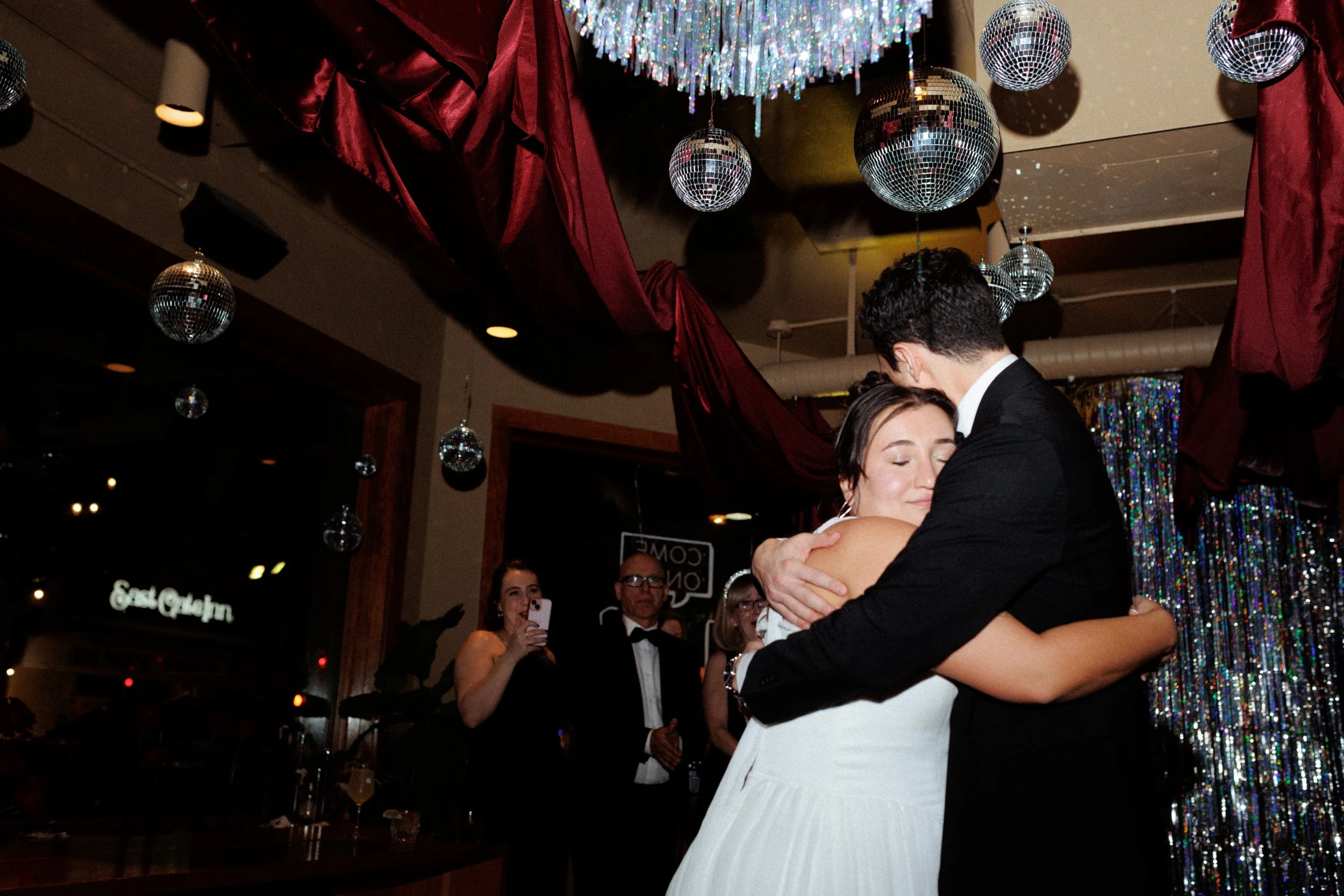A couple is embracing and sharing a kiss during a wedding reception, with guests in tuxedos and dresses watching and taking photos in the background. The setting features disco balls and festive decorations.