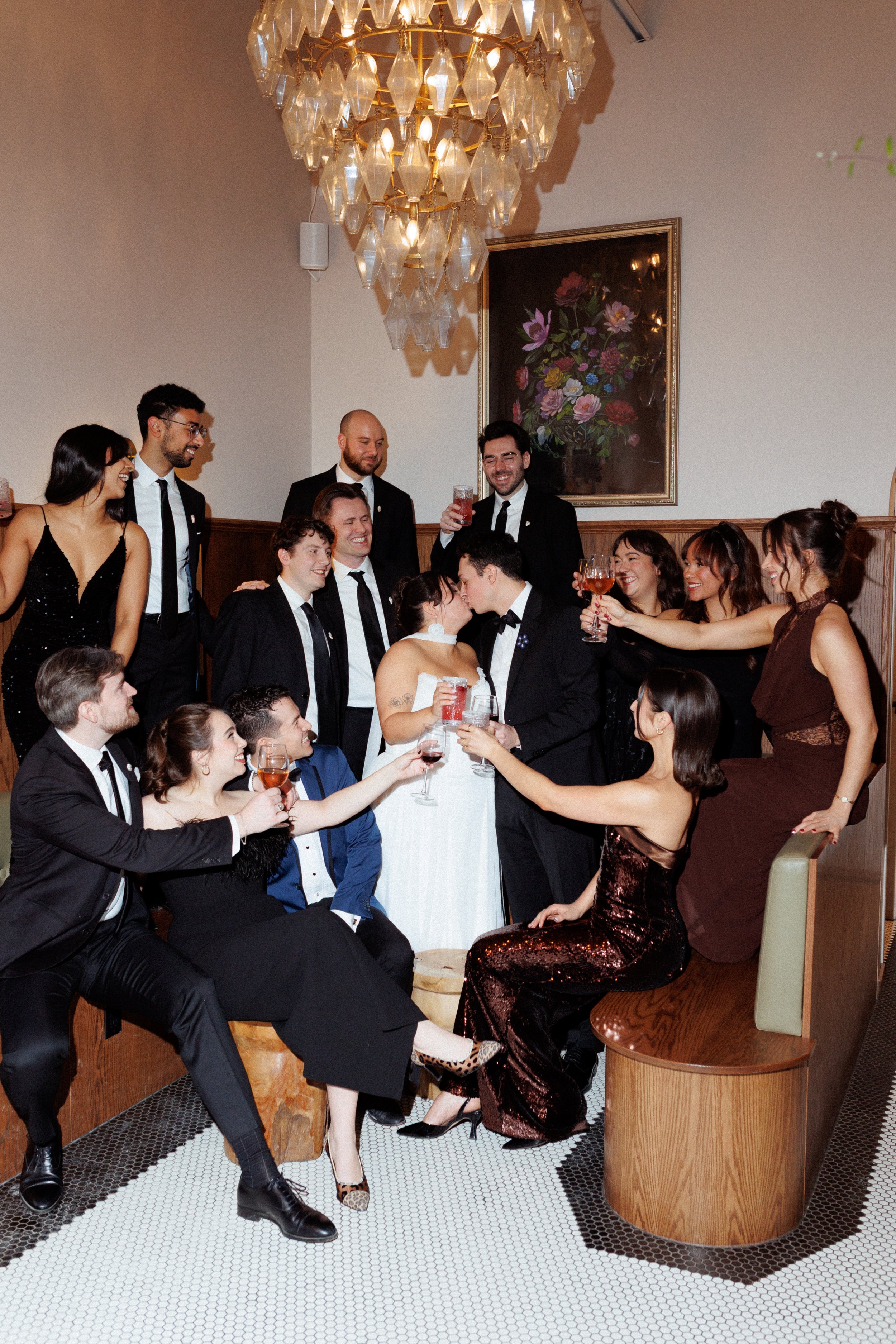 A group of people celebrating a wedding, with a bride and groom kissing, surrounded by friends raising glasses in toast, in an elegant indoor setting with a chandelier and floral artwork.