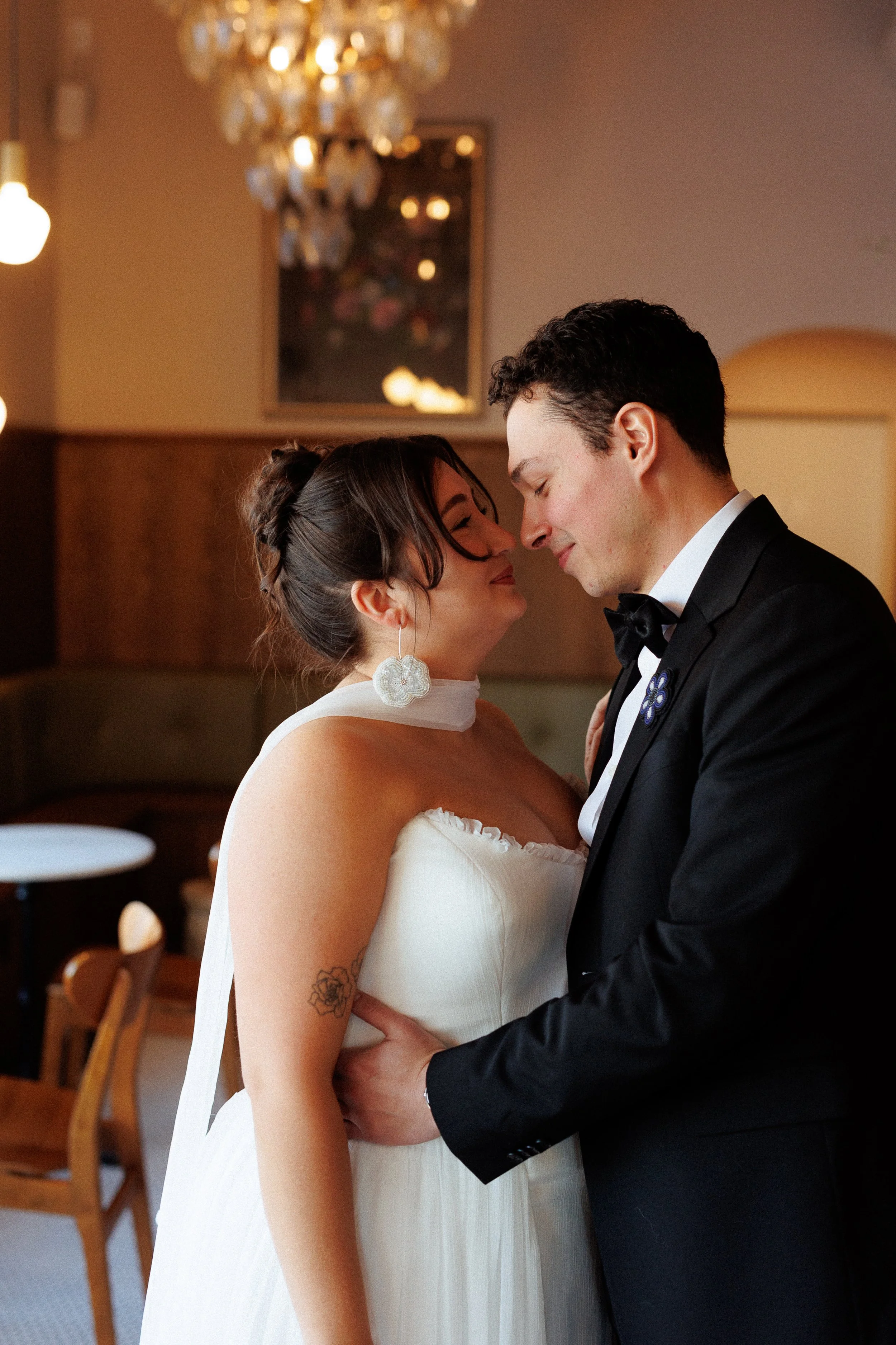 A bride and groom in wedding attire sharing an intimate moment indoors, with the bride wearing a strapless white dress and the groom in a tuxedo, their foreheads touching.