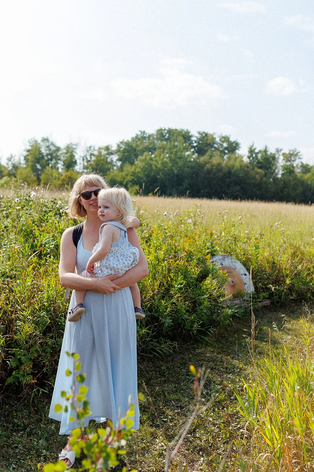 A woman in a white dress and sunglasses holding a young girl in a white dress with blue patterns in a grassy field on a sunny day.
