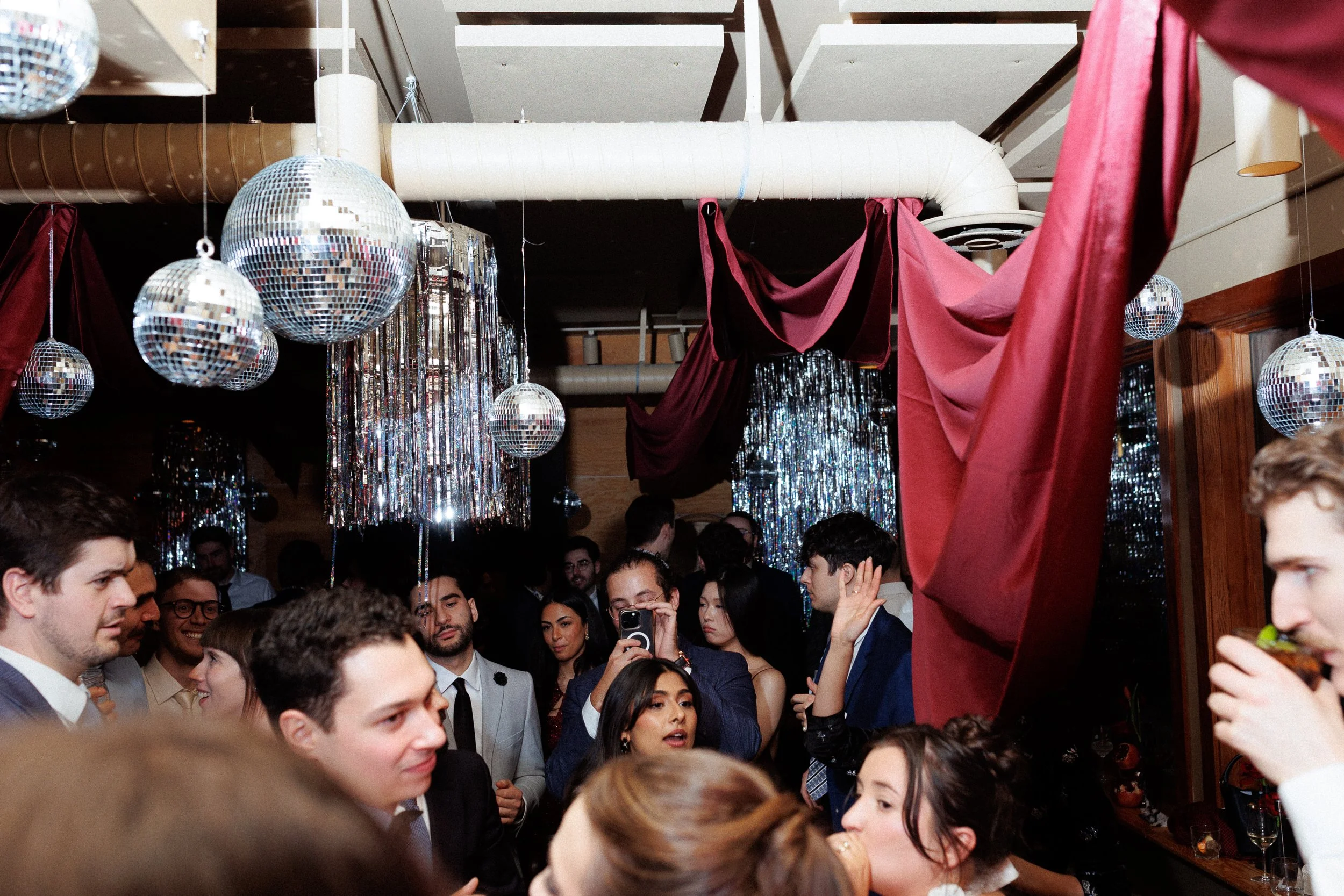 A crowded party or celebration with people talking and taking photos, decorated with disco balls, shiny streamers, and draped fabric.