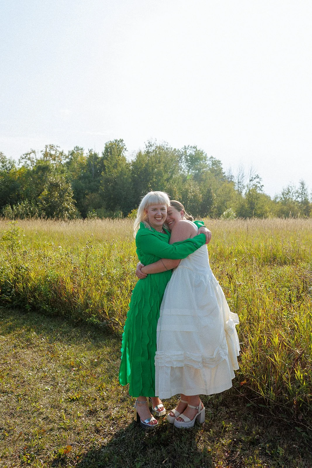 Two women hugging each other and smiling in a grassy field with trees in the background, one wearing a green dress and the other wearing a white dress.