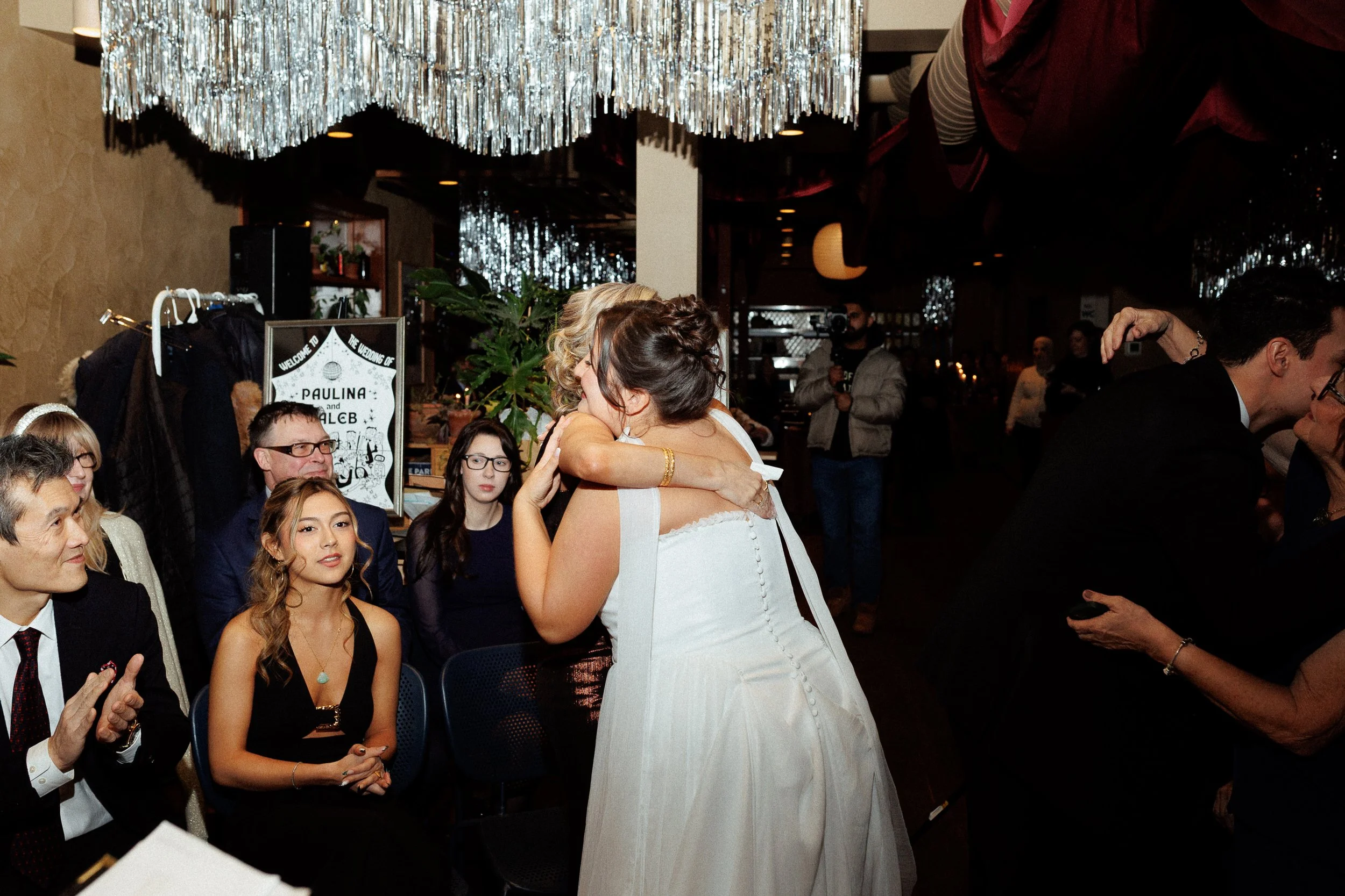 Two women embracing and kissing at a wedding reception surrounded by seated guests, with decorations and a sign in the background.