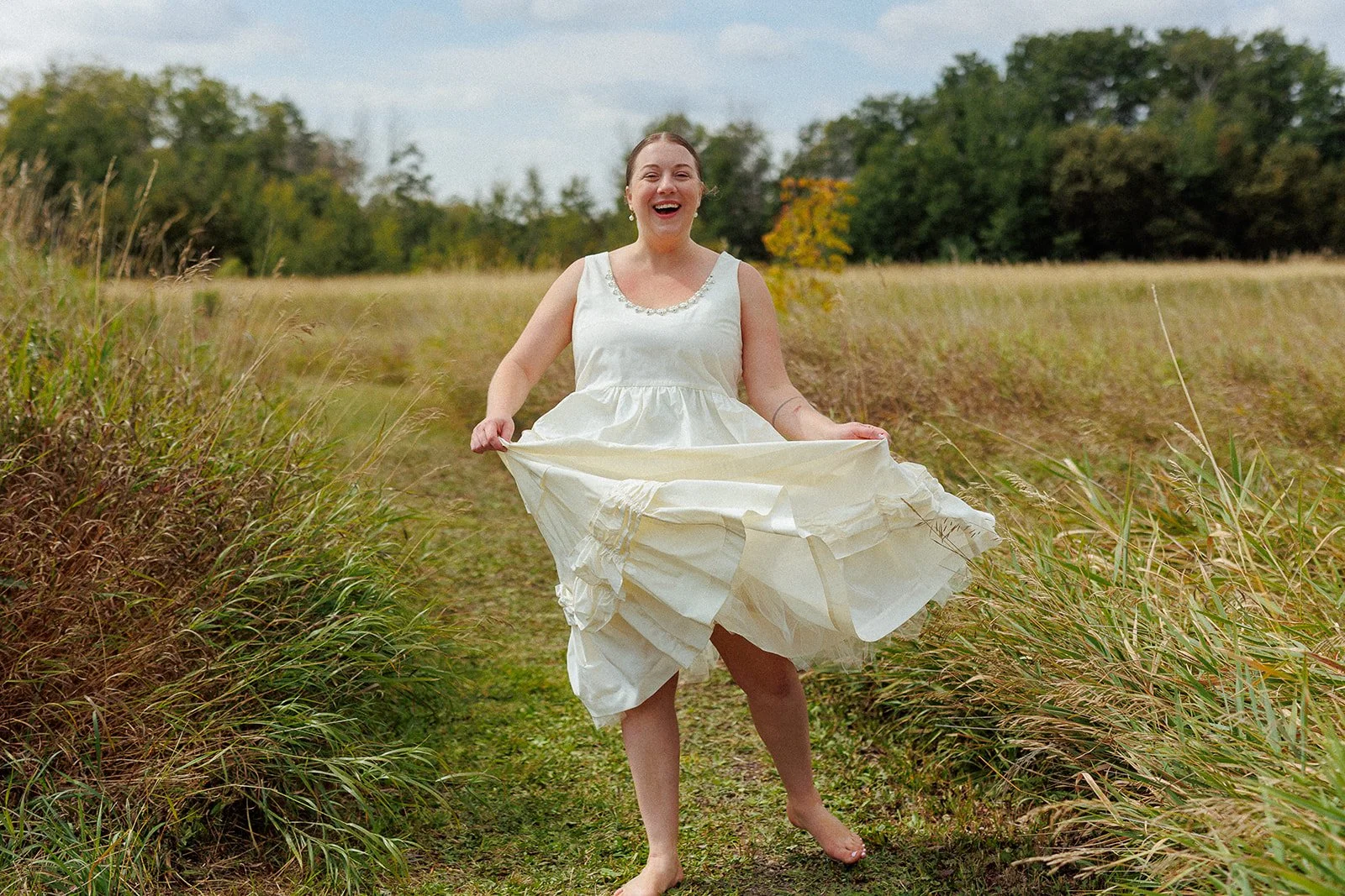 A woman in a white dress standing barefoot in a grassy field, smiling and lifting her dress while the wind blows.