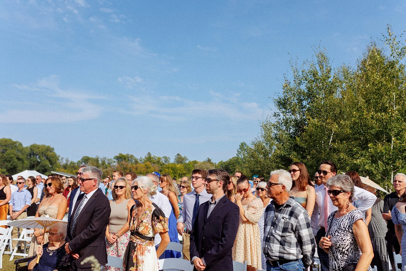 Crowd of people attending an outdoor event on a sunny day, standing and sitting under trees, with blue sky and clouds in the background.