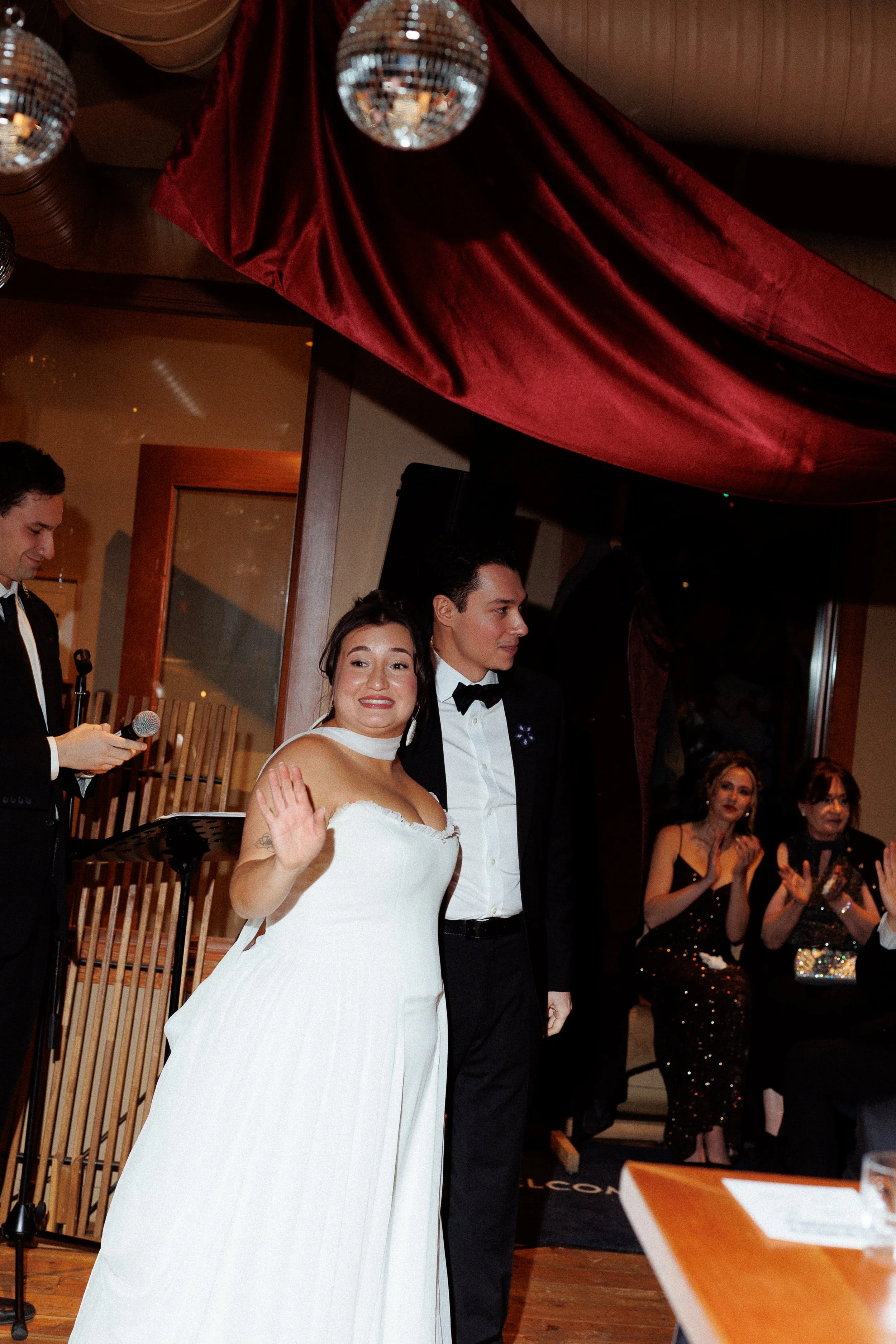 A bride and groom standing together at their wedding reception, with the bride waving and smiling, surrounded by guests clapping and celebrating.