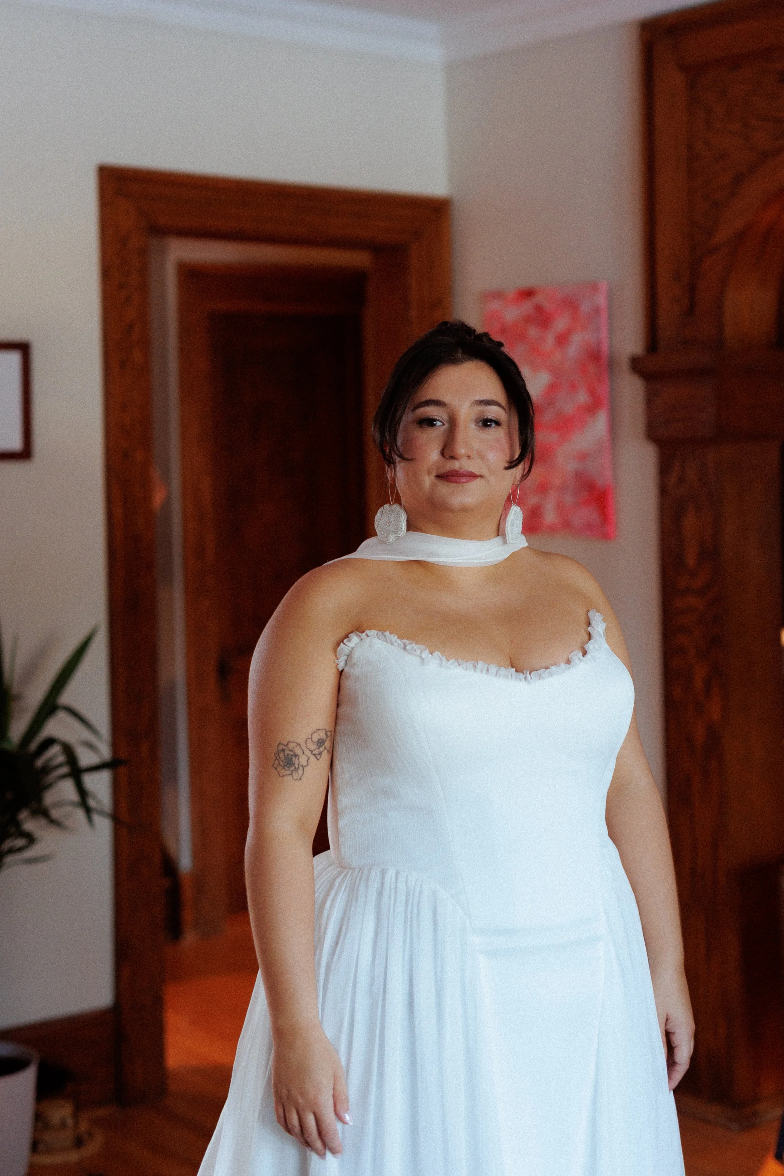 A woman in a white dress stands indoors with wooden furnishings and a pink painting in the background.