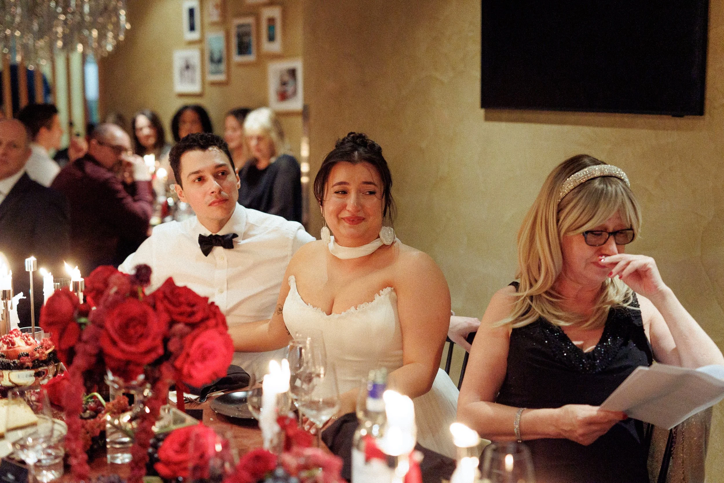A woman in a white dress with a choker and large earrings sitting at a table, smiling. A man in a white shirt with a bow tie sitting next to her. An older woman in a black dress with glasses reading a program or menu. Several people are visible in th