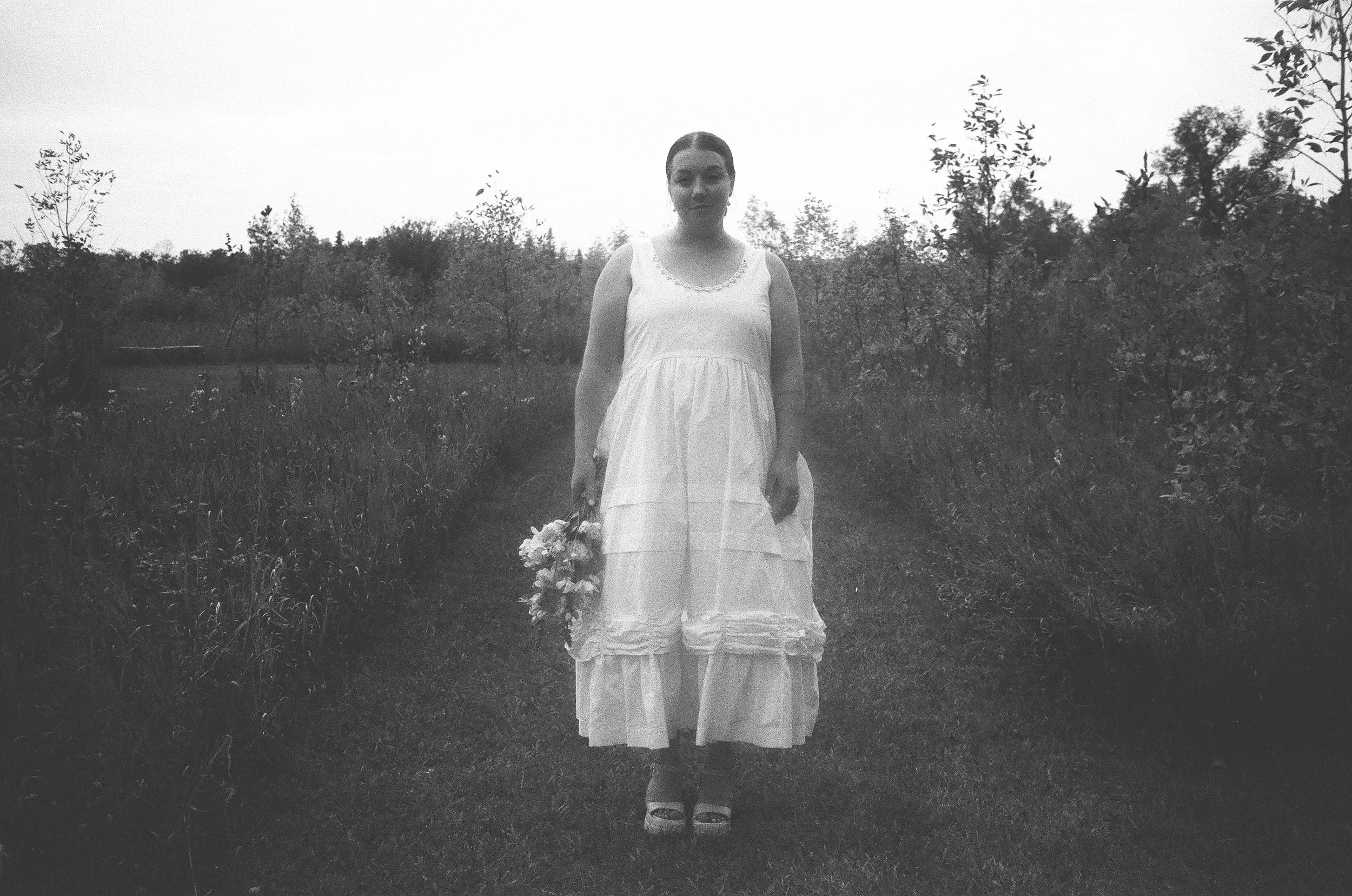 A woman in a long, flowing white dress standing outdoors on a grassy path, holding a bouquet of flowers in her left hand, with trees and shrubs in the background.