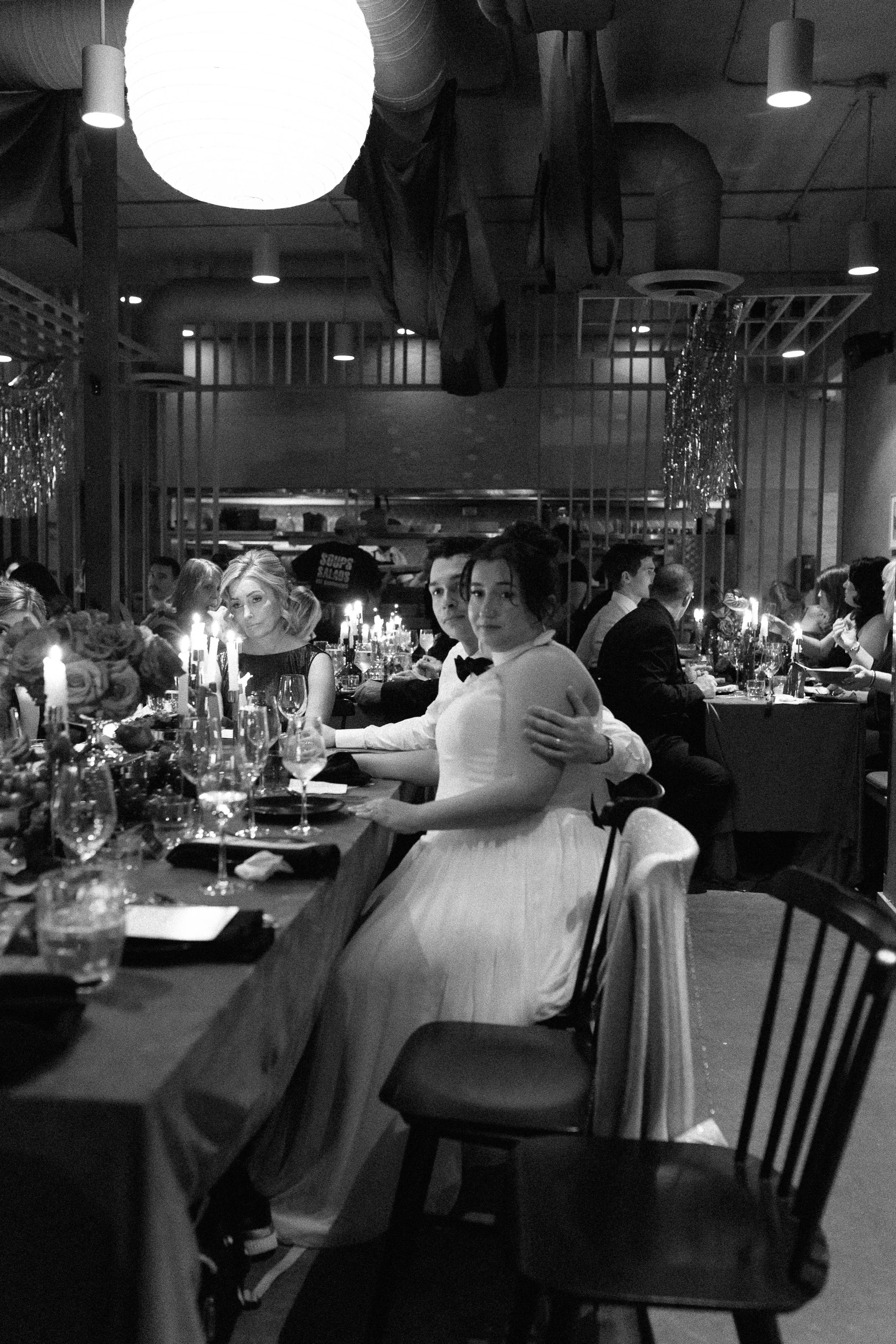 People sitting at a dinner table in a dimly lit restaurant, including a woman in a white dress and a man in a tuxedo, with candles and flower arrangements on the table.
