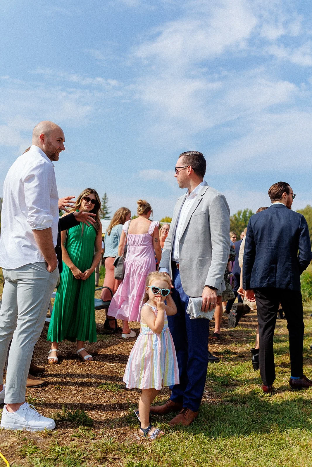 Group of people outdoor on a sunny day, including a man in a grey blazer, a girl with sunglasses, and various women dressed in colorful attire.