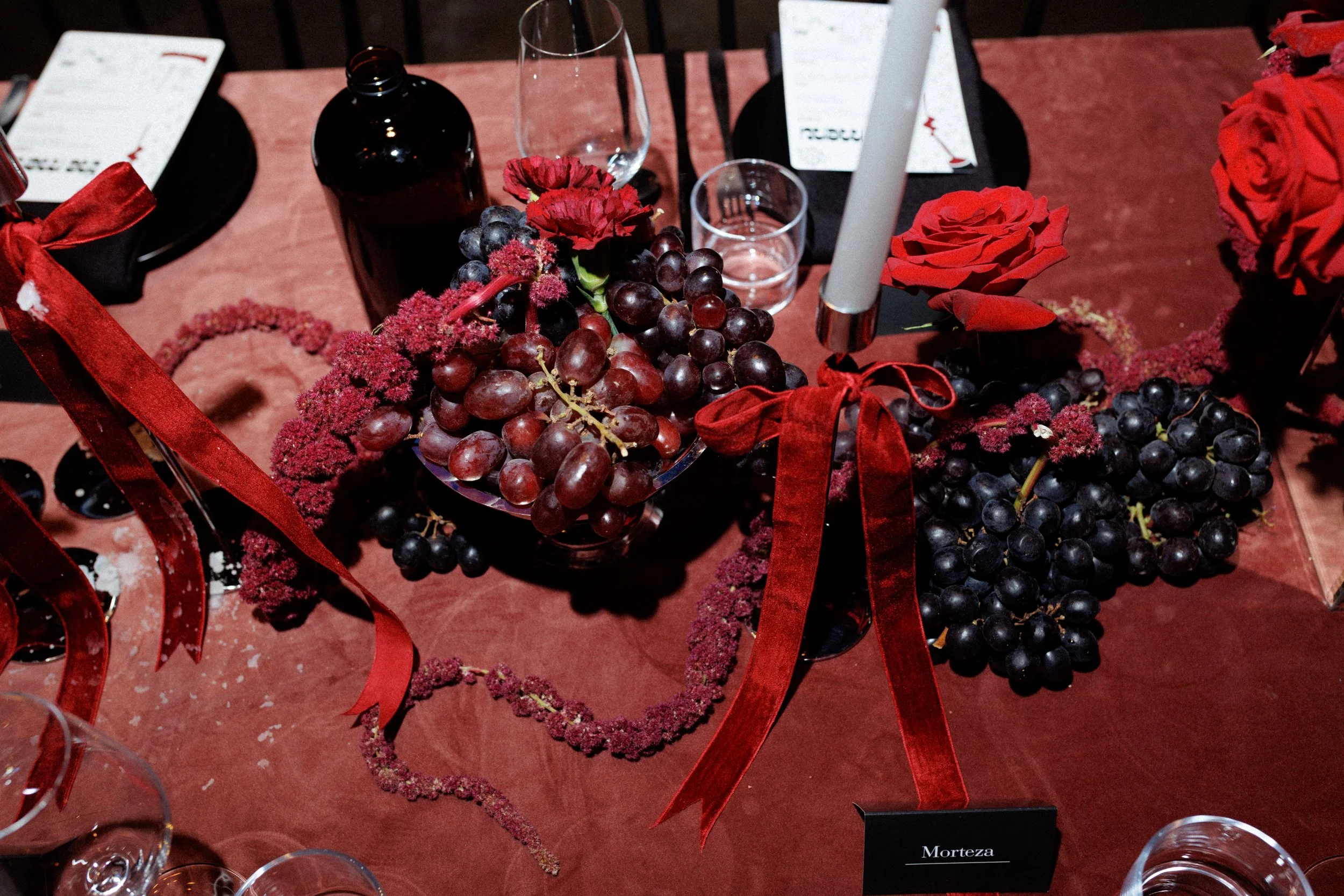 A dinner table decorated with bunches of dark red and purple grapes, red roses, and red ribbons, along with a white candle, a wine glass, and a black bottle.
