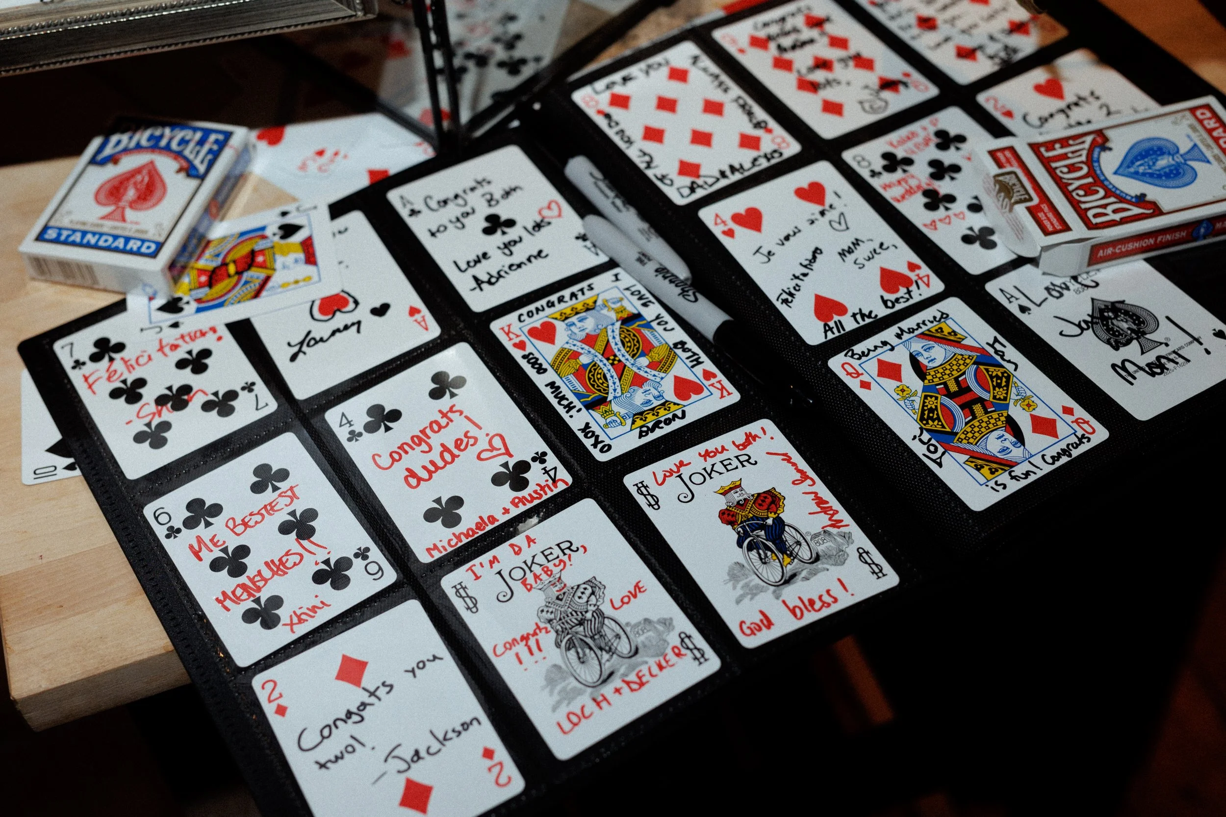 Photograph of a playing card collection in a binder, with handwritten notes and messages in red ink, including congratulatory notes and personal messages, along with decks of Bicycle playing cards on a wooden surface.