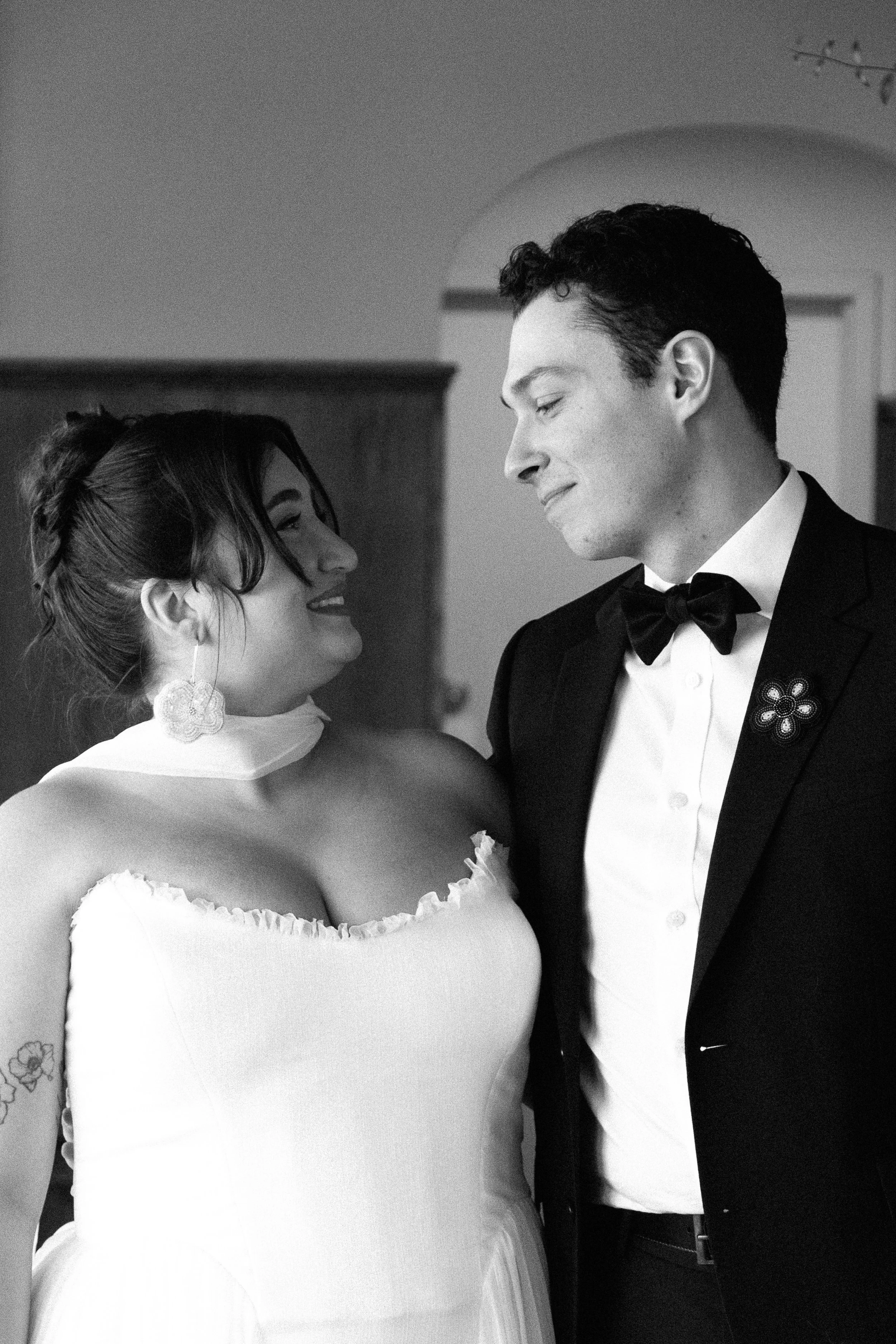 A black and white photo of a bride and groom smiling at each other indoors. The bride wears a strapless dress and floral earrings. The groom wears a tuxedo with a bow tie.