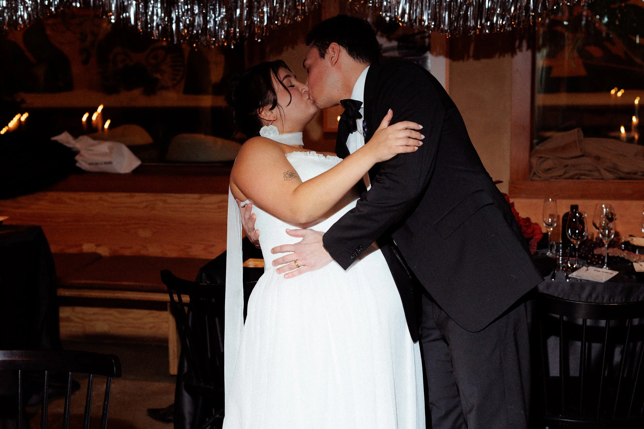 A couple dressed in wedding attire sharing a kiss, with the bride in a white gown and the groom in a black tuxedo, during a wedding reception.