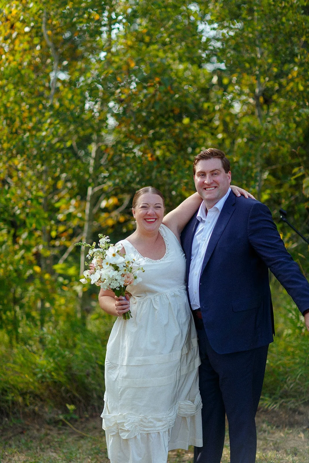 A smiling couple, the woman in a white dress holding a bouquet of flowers, and the man in a dark suit, standing outdoors in front of green trees.