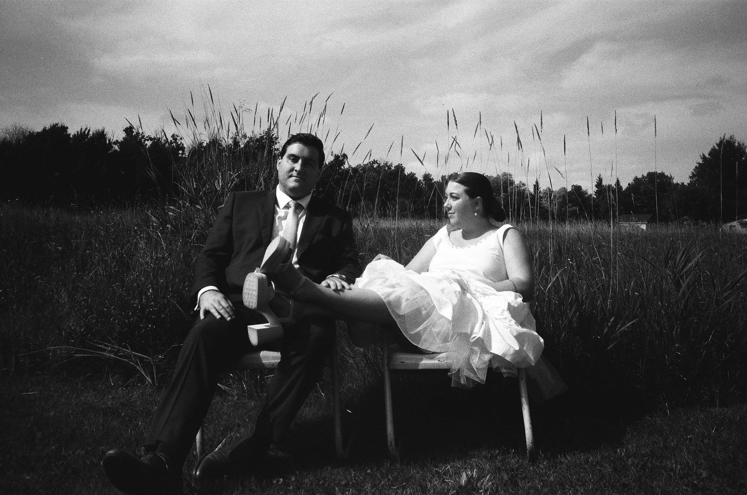 A black and white photo of a man and woman outdoors in a grassy field. The man, in a suit, is sitting on a chair and the woman, dressed in a light-colored dress, is lying on a bench with her legs resting on the man's lap.