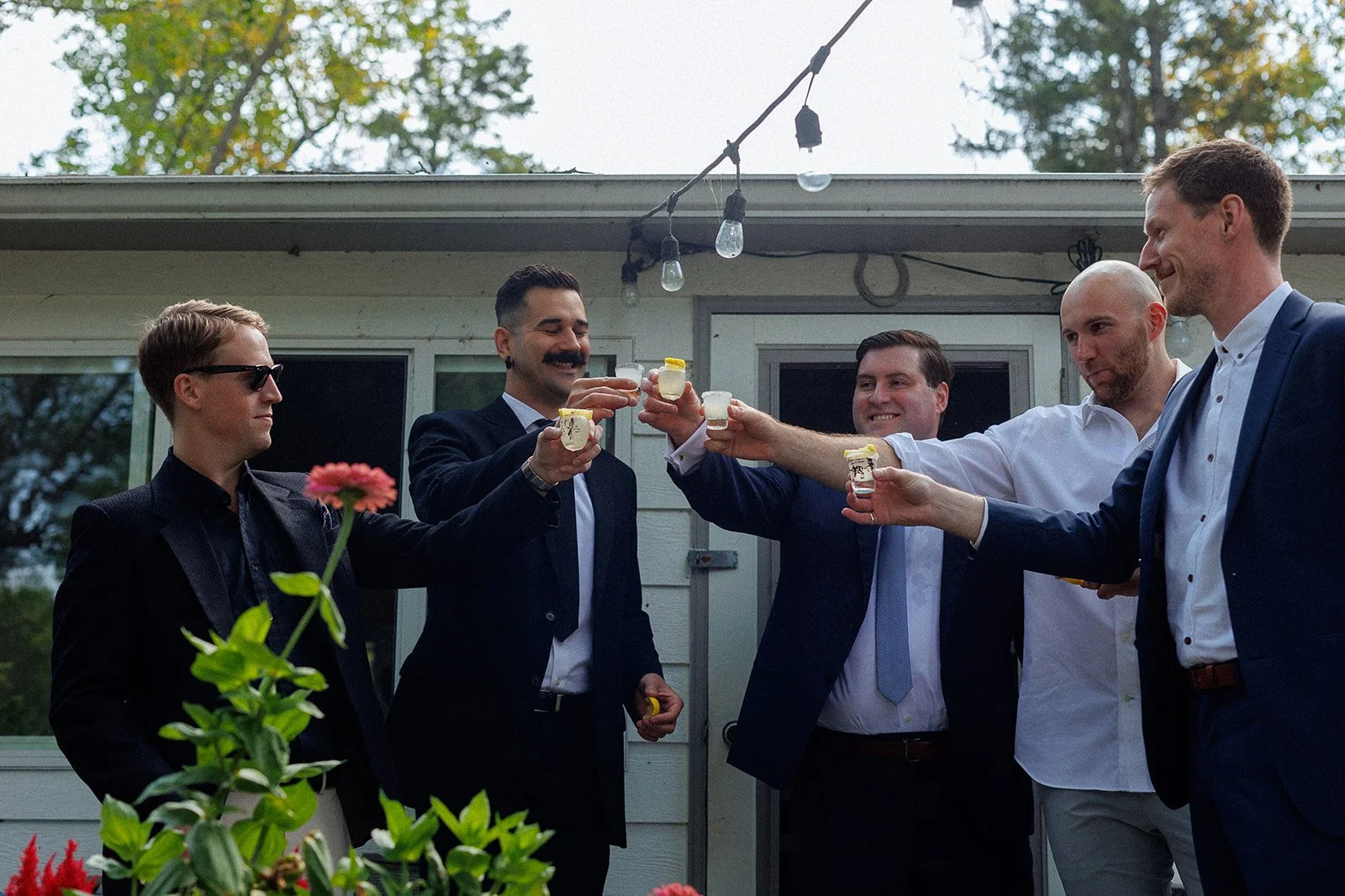 A group of five men in suits cheer and toast drinks outdoors during a celebration, with a house and trees in the background.