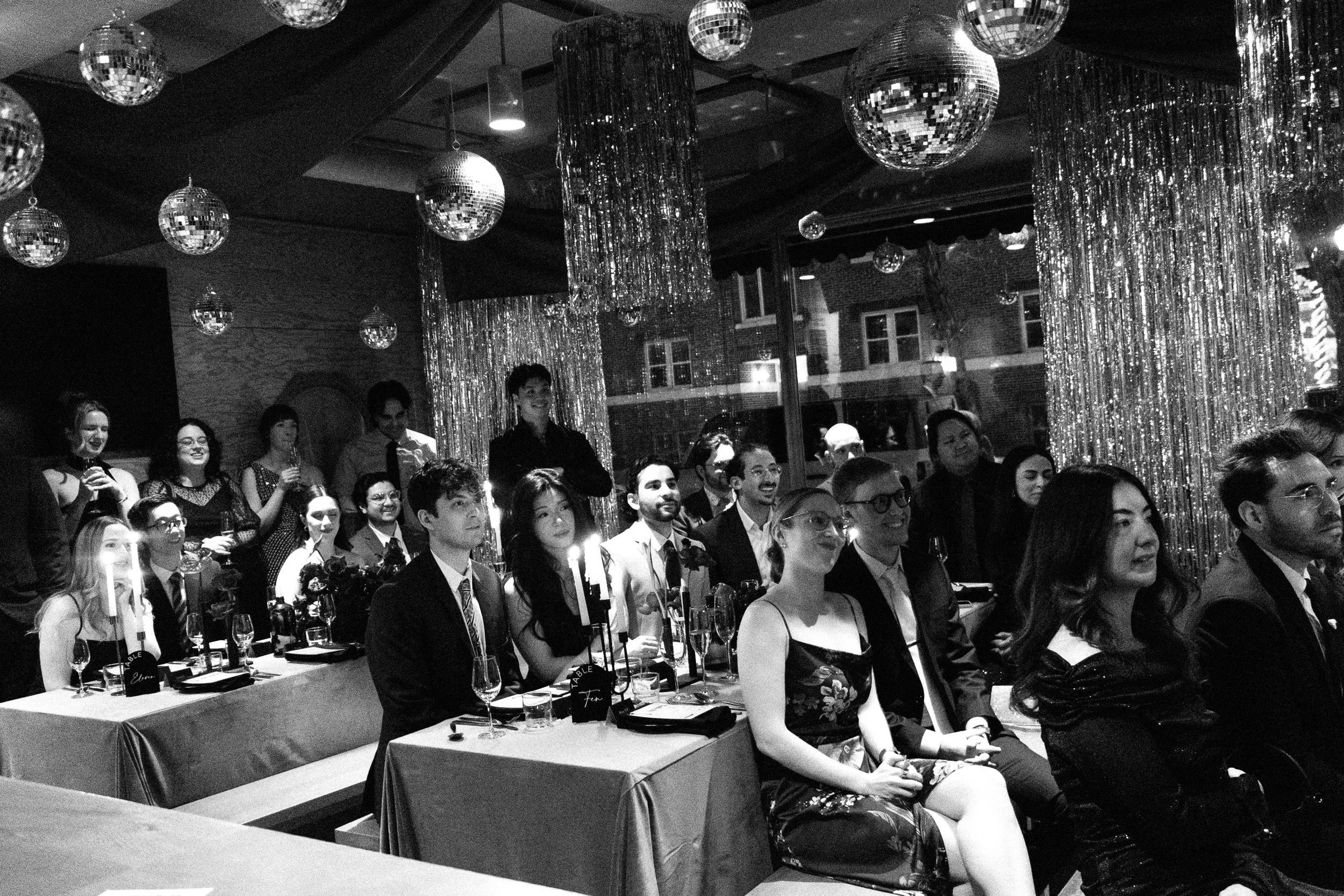 A group of people attending a formal event or celebration, seated at tables with glasses and candles, in a venue decorated with shiny hanging disco balls and curtains.