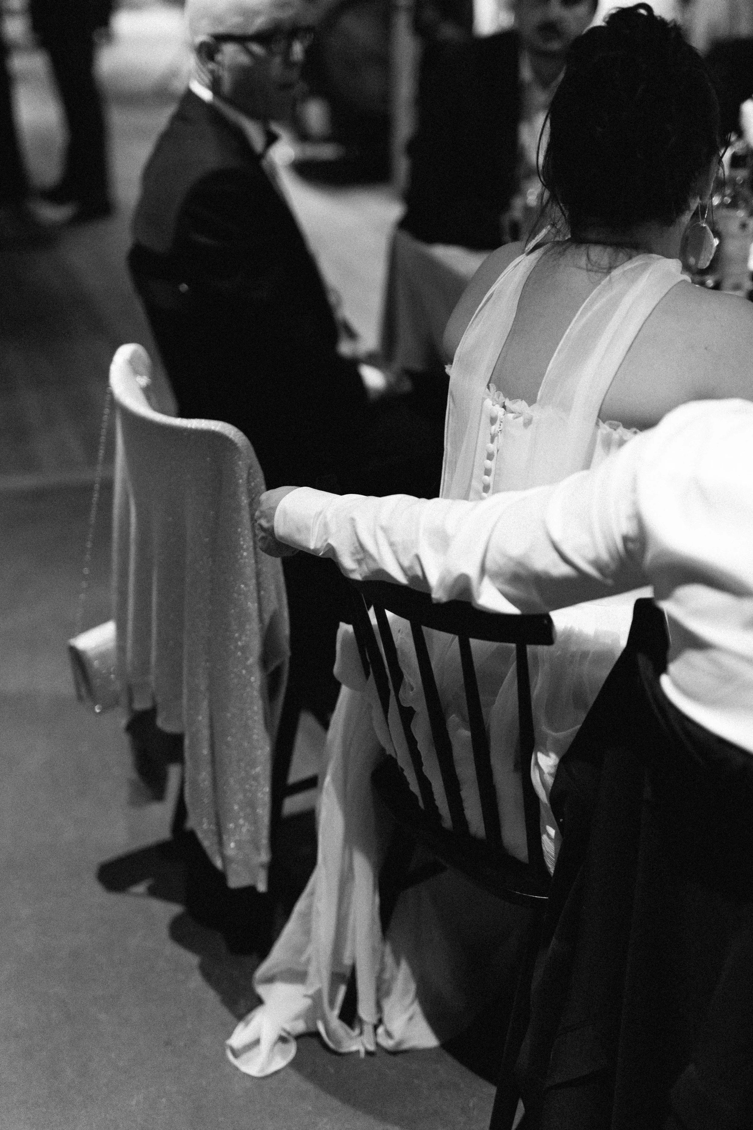 Black and white photo of people sitting at a table during a formal event, with a woman in a light-colored dress and a man in a white shirt and a tuxedo.