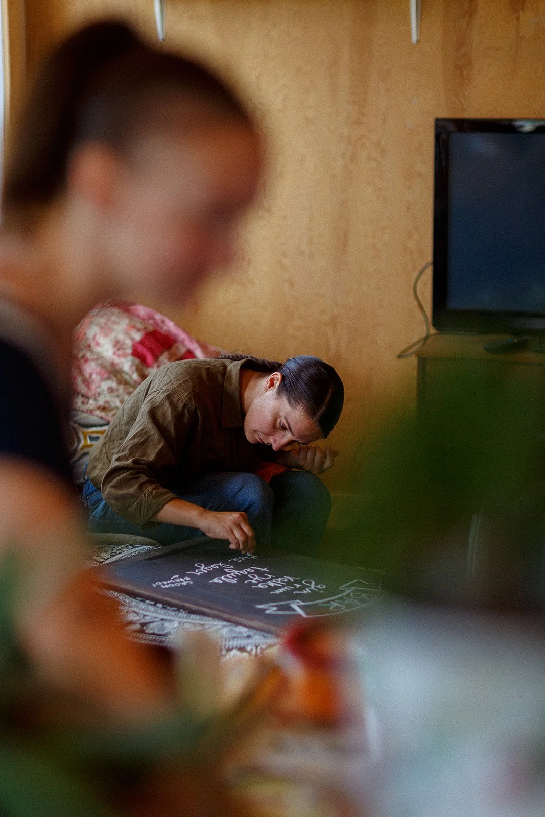 A woman sitting on a couch writing on a blackboard with white chalk, with another person blurred in the foreground and a television on a stand in the background.
