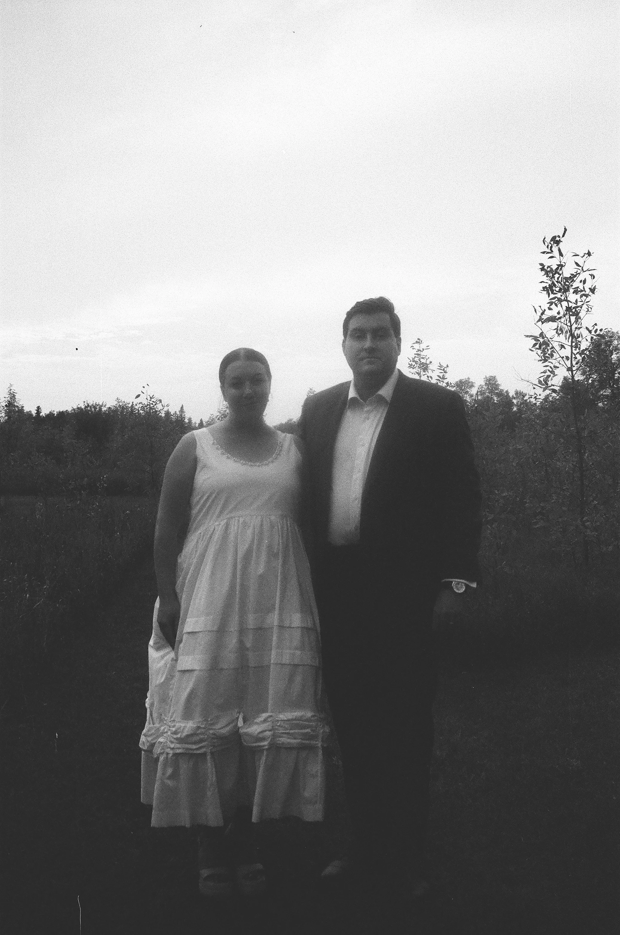 A man and woman standing outdoors in a grassy area with trees in the background, dressed in formal attire, with the woman wearing a long white dress and the man wearing a dark suit and tie.