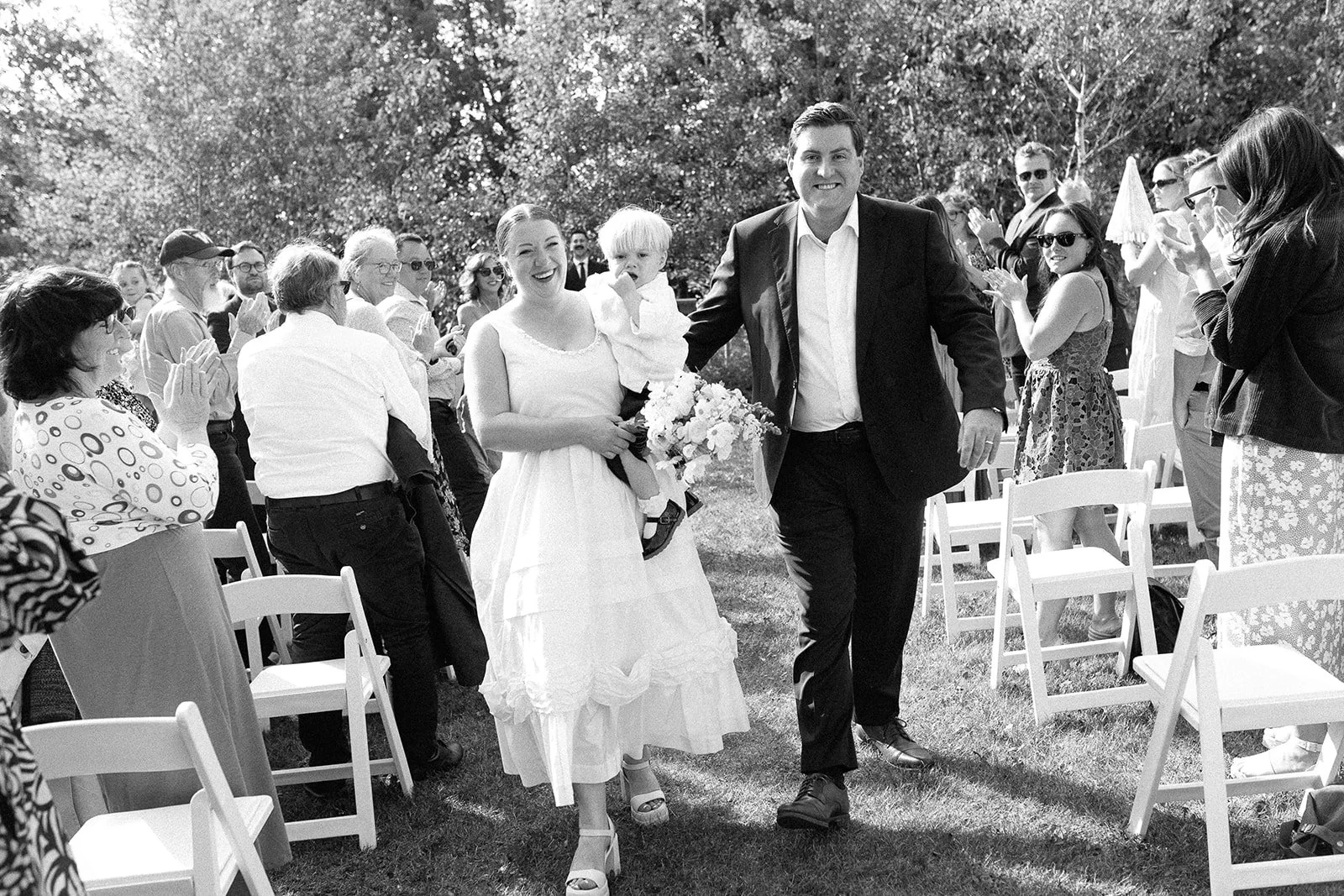 A bride and groom walking down the aisle at an outdoor wedding, smiling and holding flowers, surrounded by seated and standing guests applauding and celebrating.
